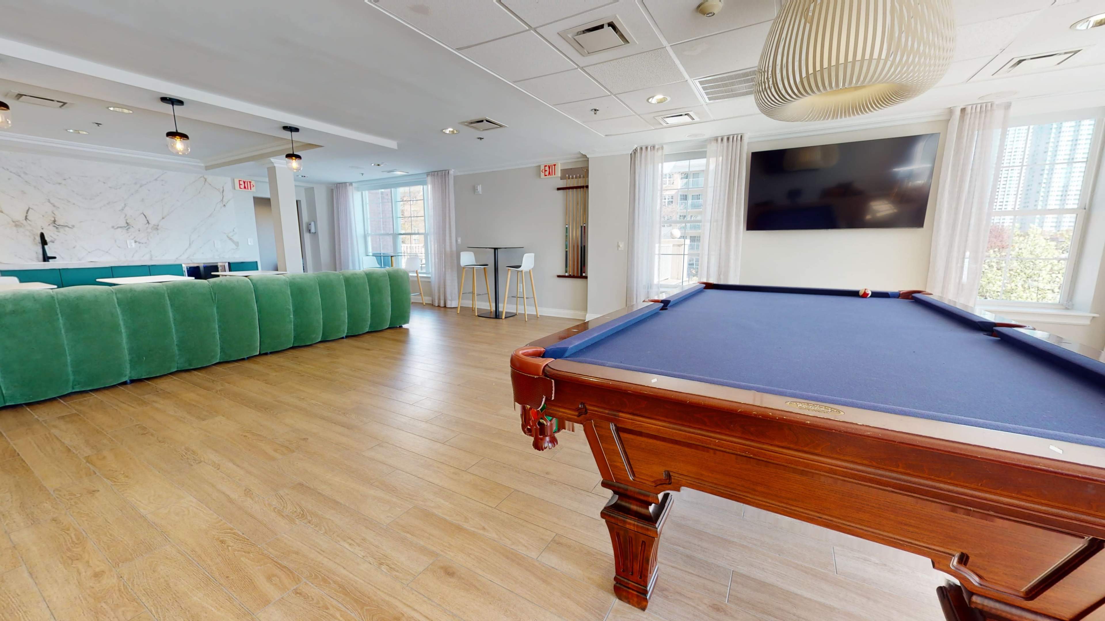 The scene features a billiards table in a modern recreational room with a green couch, a large wall-mounted TV, and sleek bar stools.