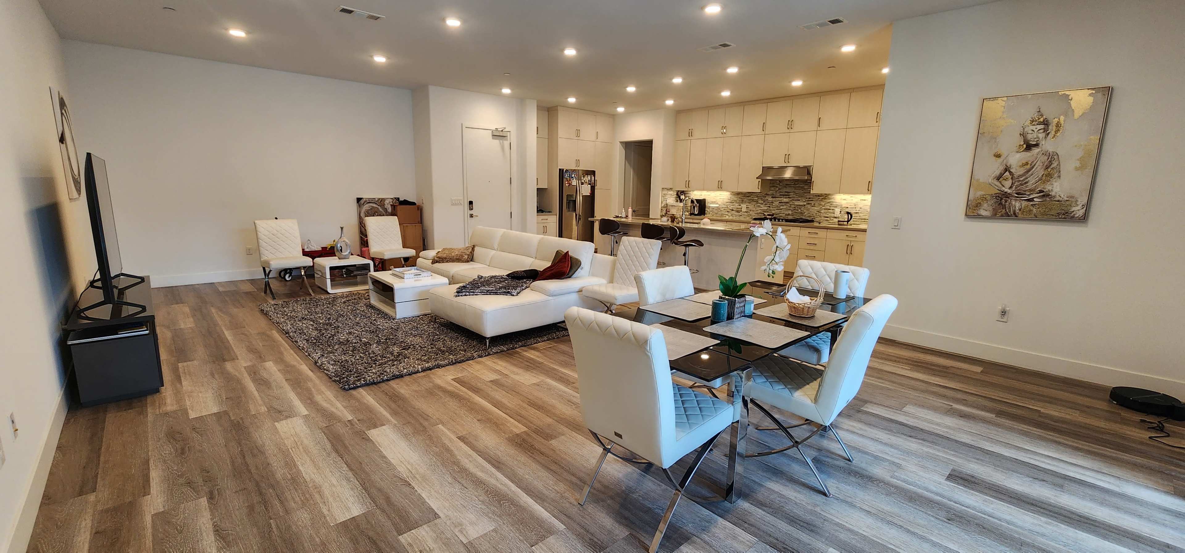A modern living room features a white sectional sofa, a glass dining table with chairs, and an open-concept kitchen in the background.
