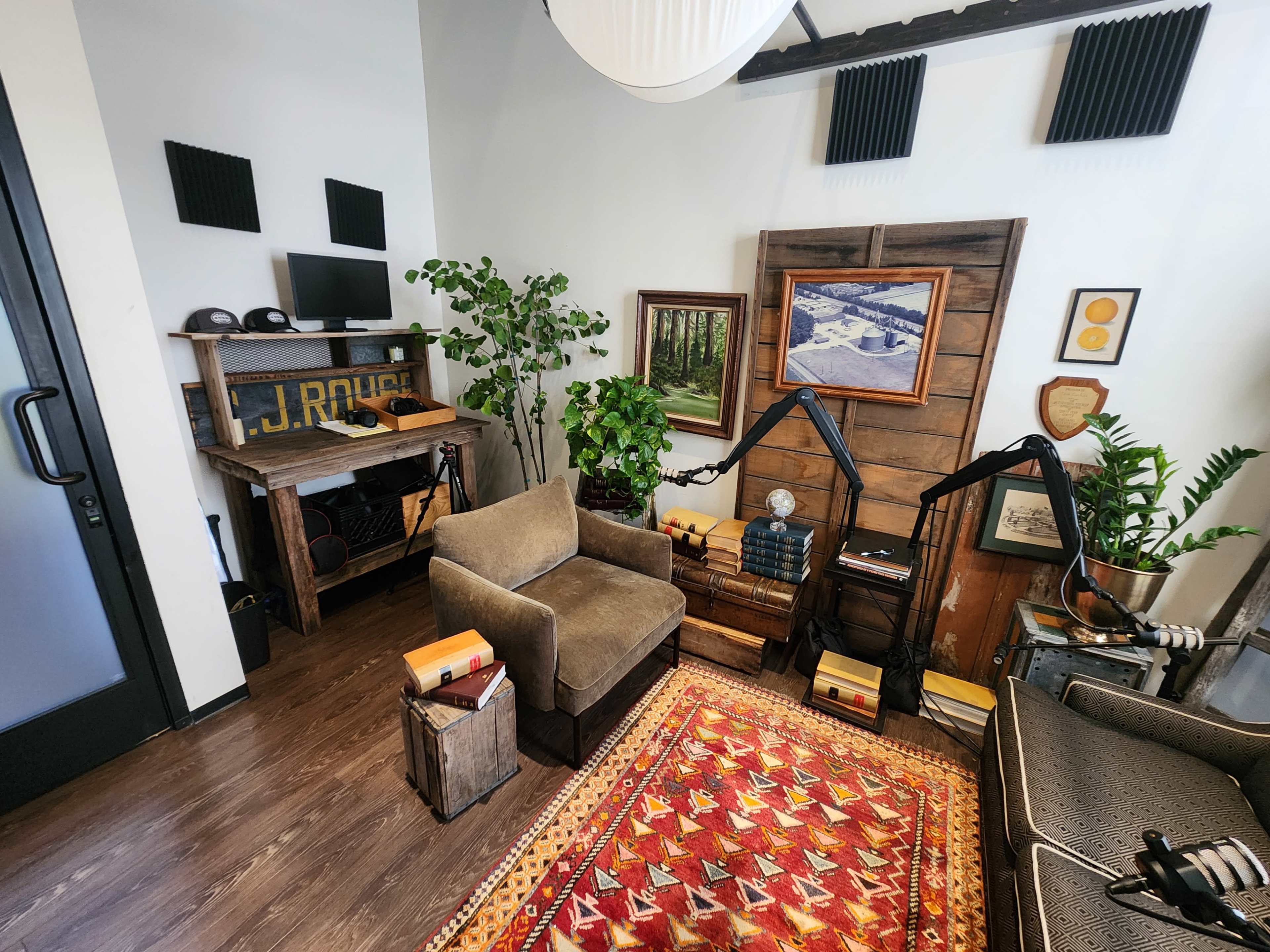 The image shows a cozy office space featuring a brown armchair, wooden desk with plants, a colorful rug, and wall décor, including a framed picture and acoustic panels.
