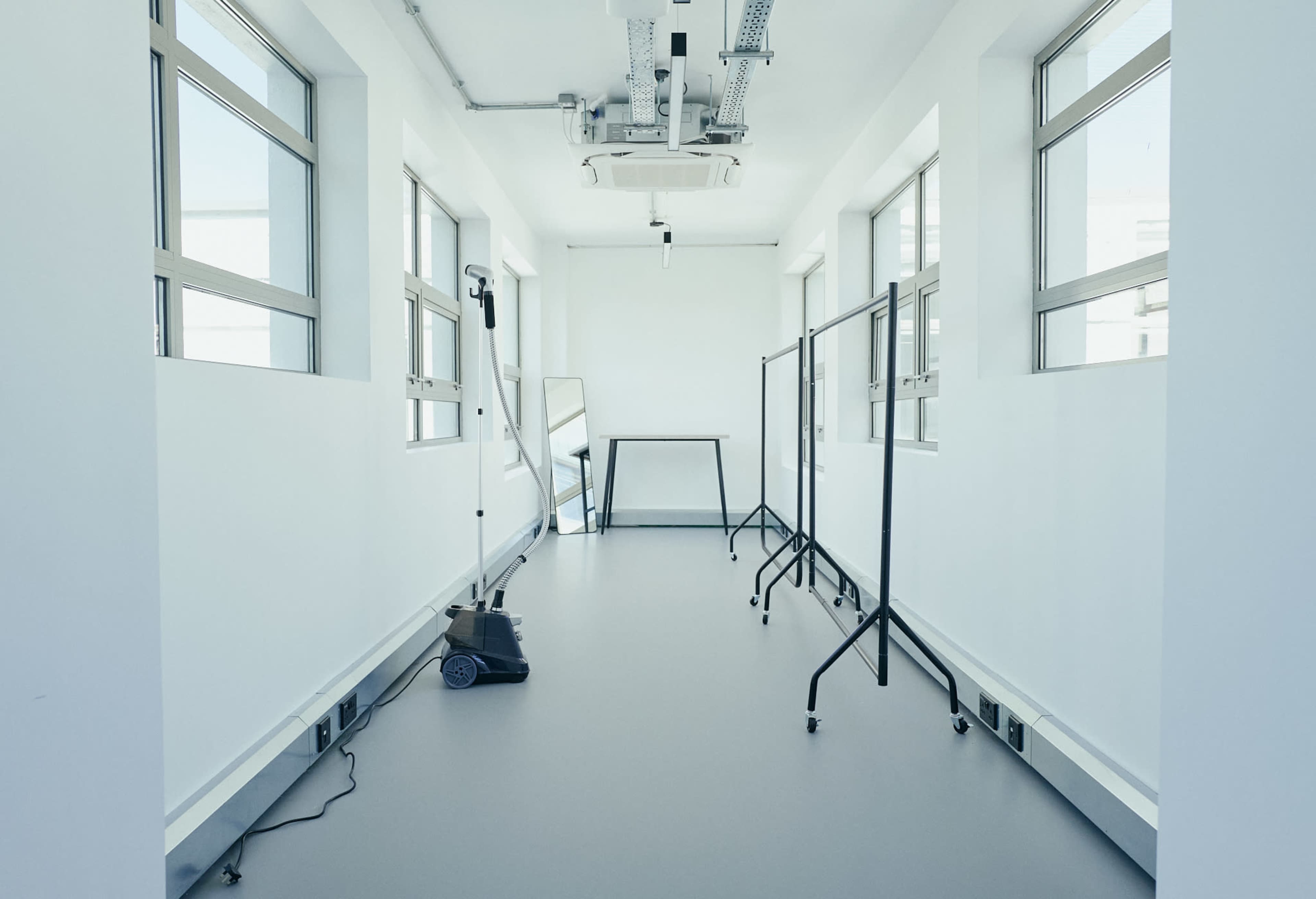 The image shows a spacious, empty room with white walls, large windows, and minimal furniture, including a mirror and some clothing racks.