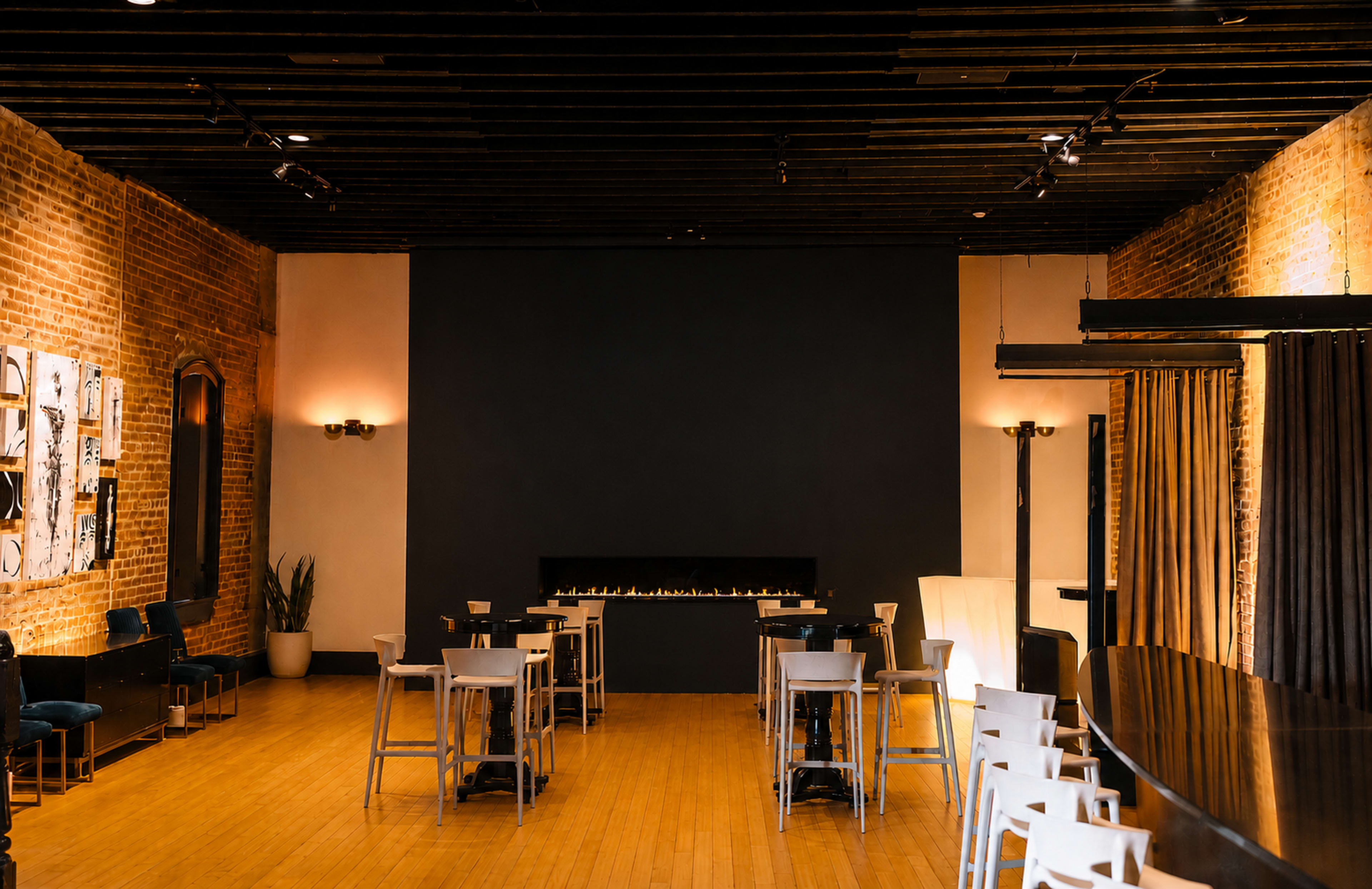 The image shows a spacious venue with wooden floors, exposed brick walls, a dark feature wall, and a long bar area with seating and a minimalist fireplace.