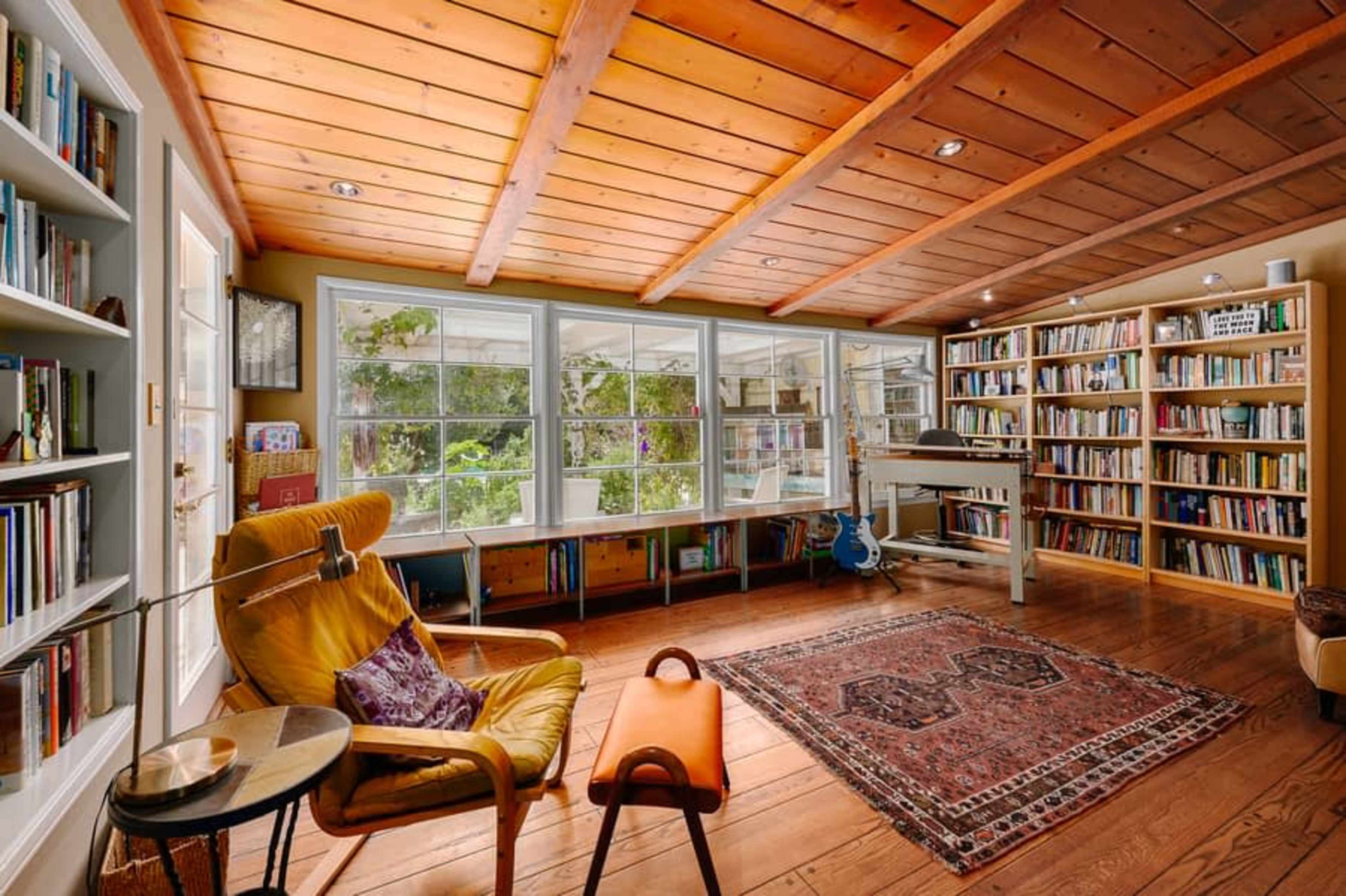 A cozy reading room features a yellow armchair and a side table, surrounded by bookshelves and large windows with a view of greenery.