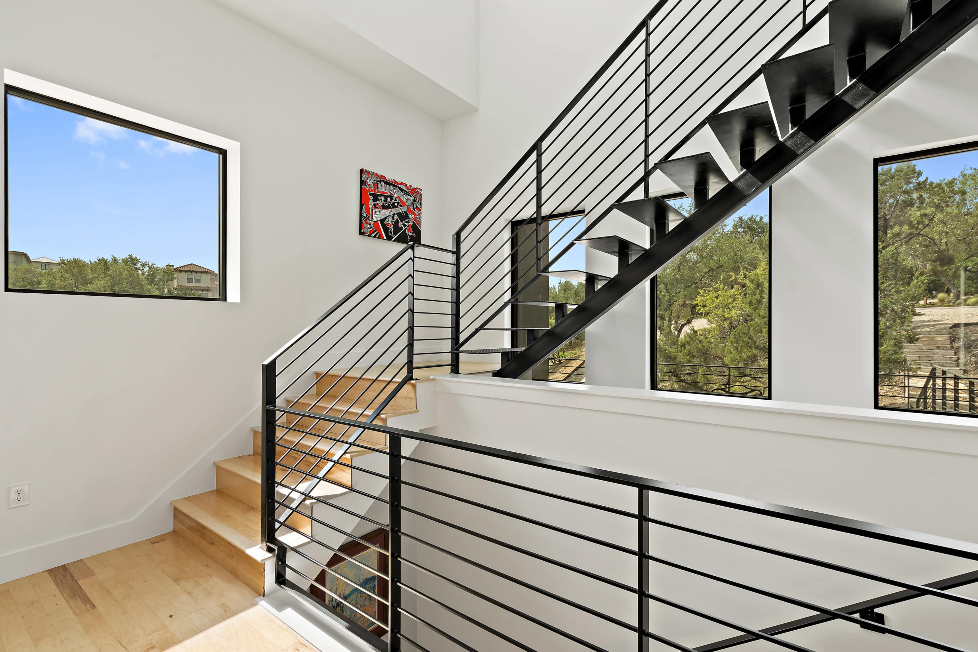 The image shows a modern staircase with black railings and wooden steps, surrounded by large windows that allow natural light to enter the space.