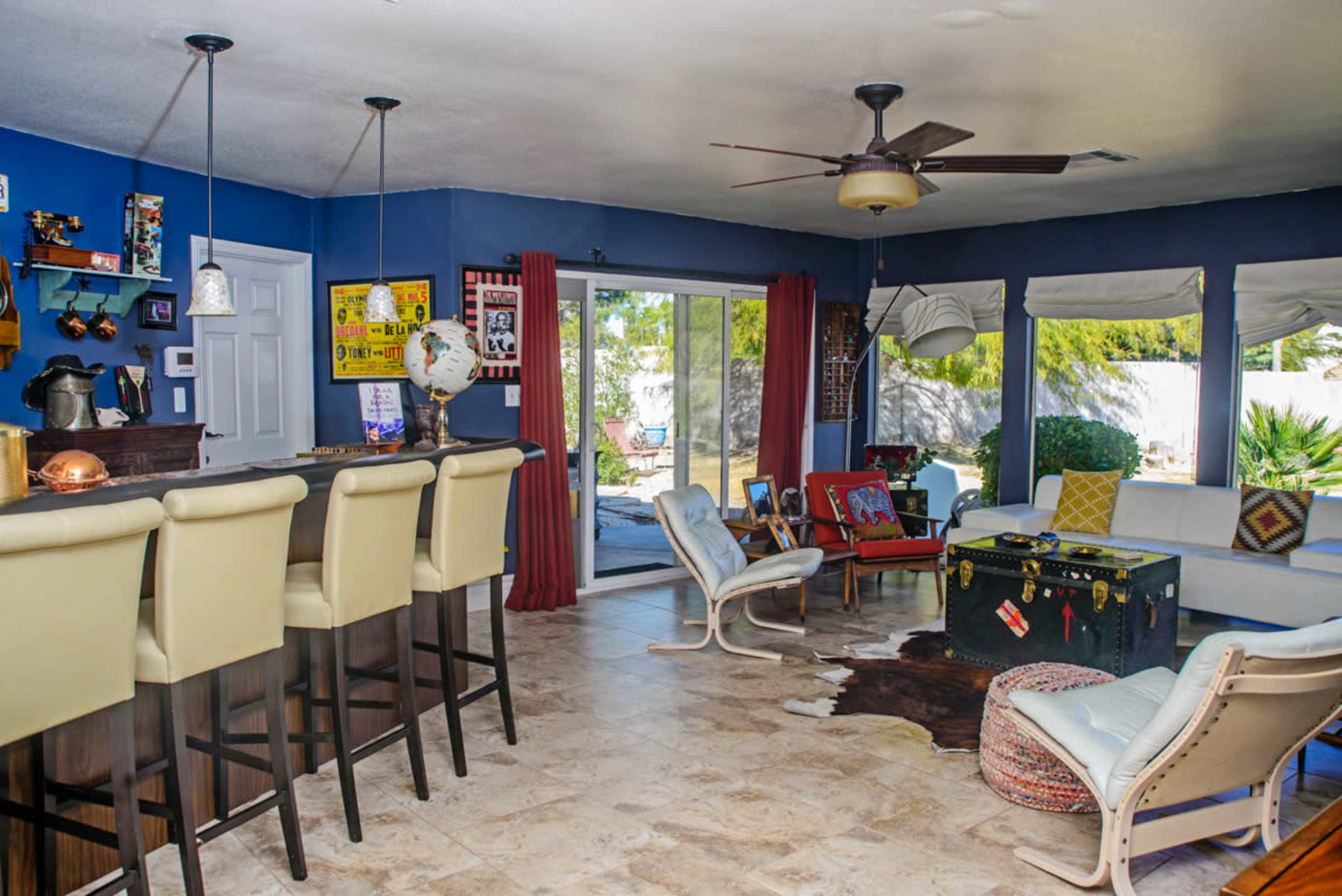 A living room with a bar area, several chairs, and a door leading to an outdoor space with a view of greenery.