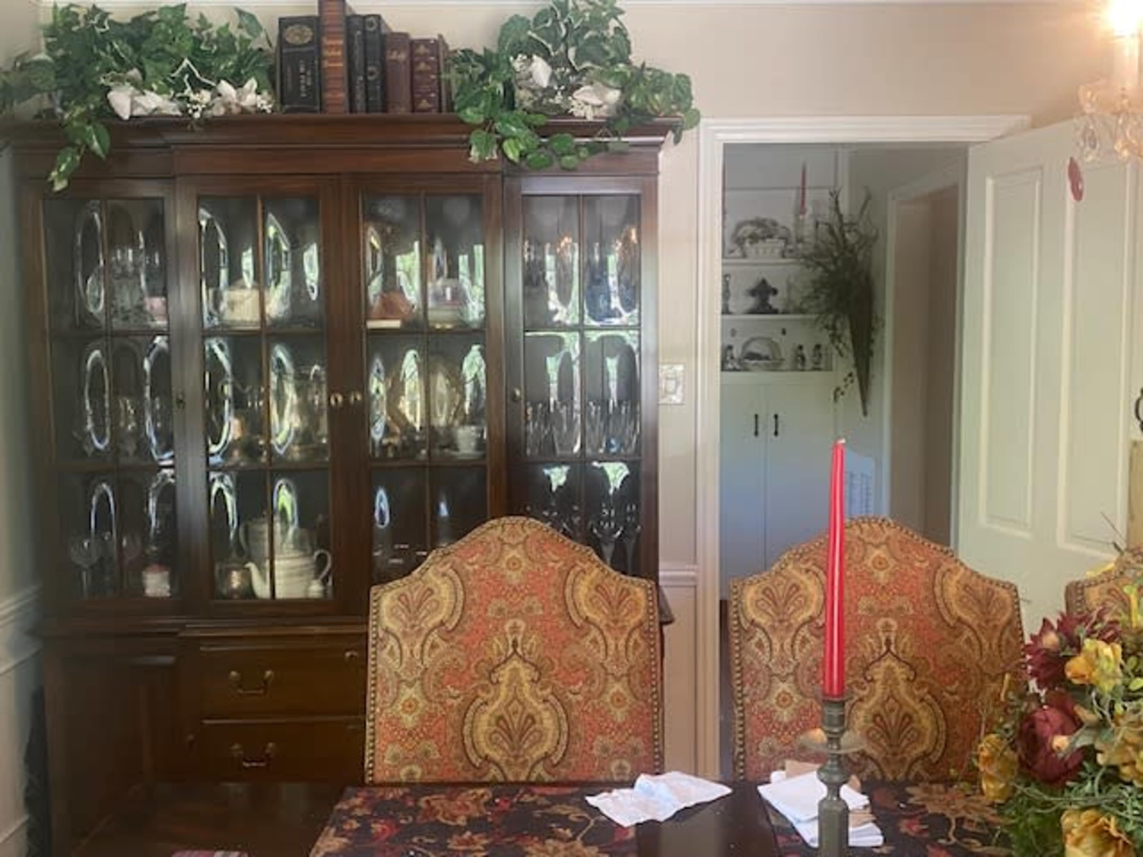 A dining room features a wooden china cabinet displaying glassware and two ornate chairs at a table with a red candle and floral centerpiece.