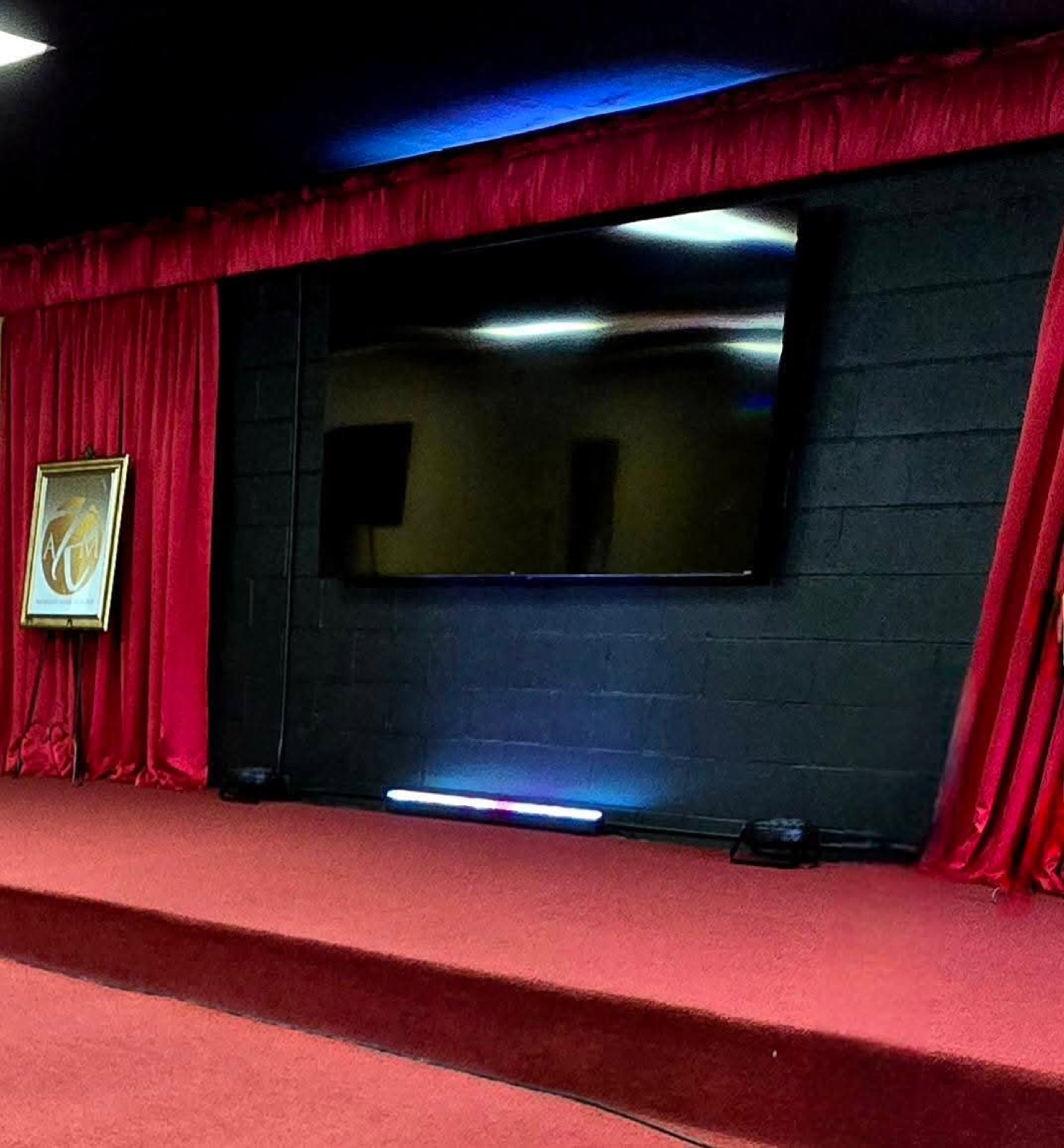 The image shows a stage with a red carpet, flanked by red curtains, and a large black screen on a black wall.