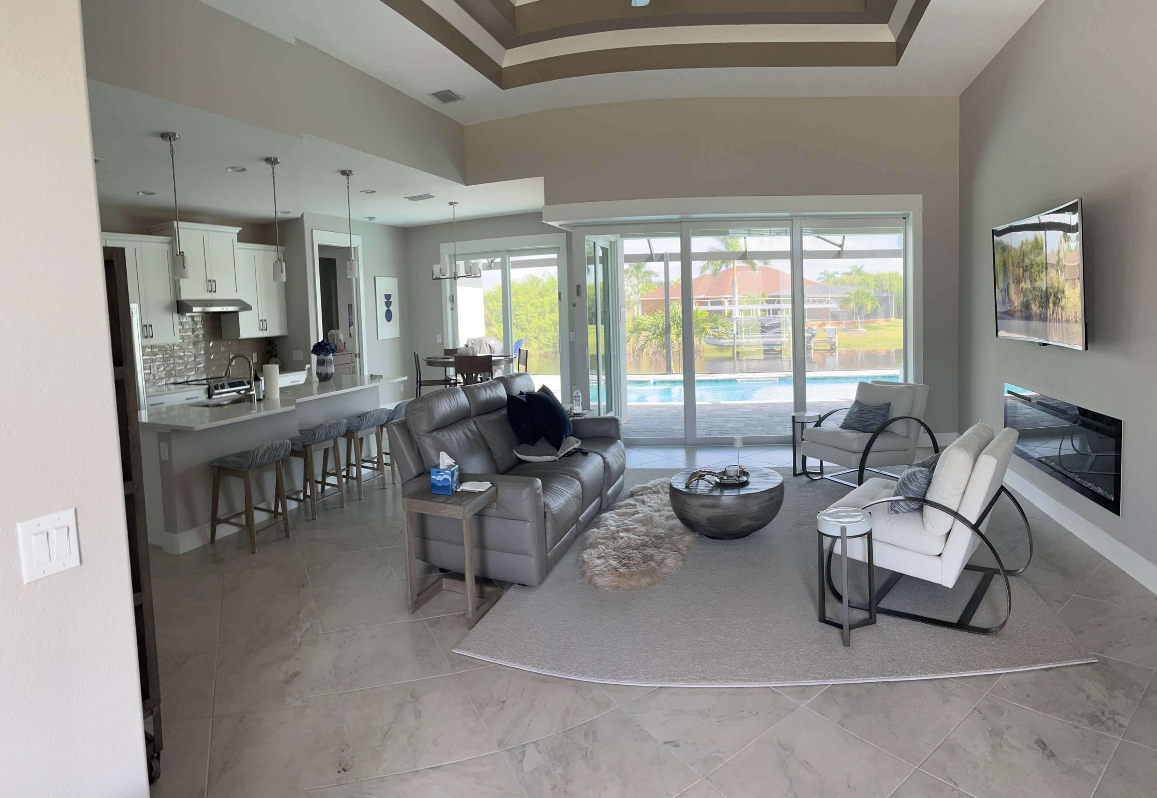 The image shows a modern living room with a gray leather sofa, a coffee table, and large glass doors leading to a pool area, flanked by a kitchen with bar seating.