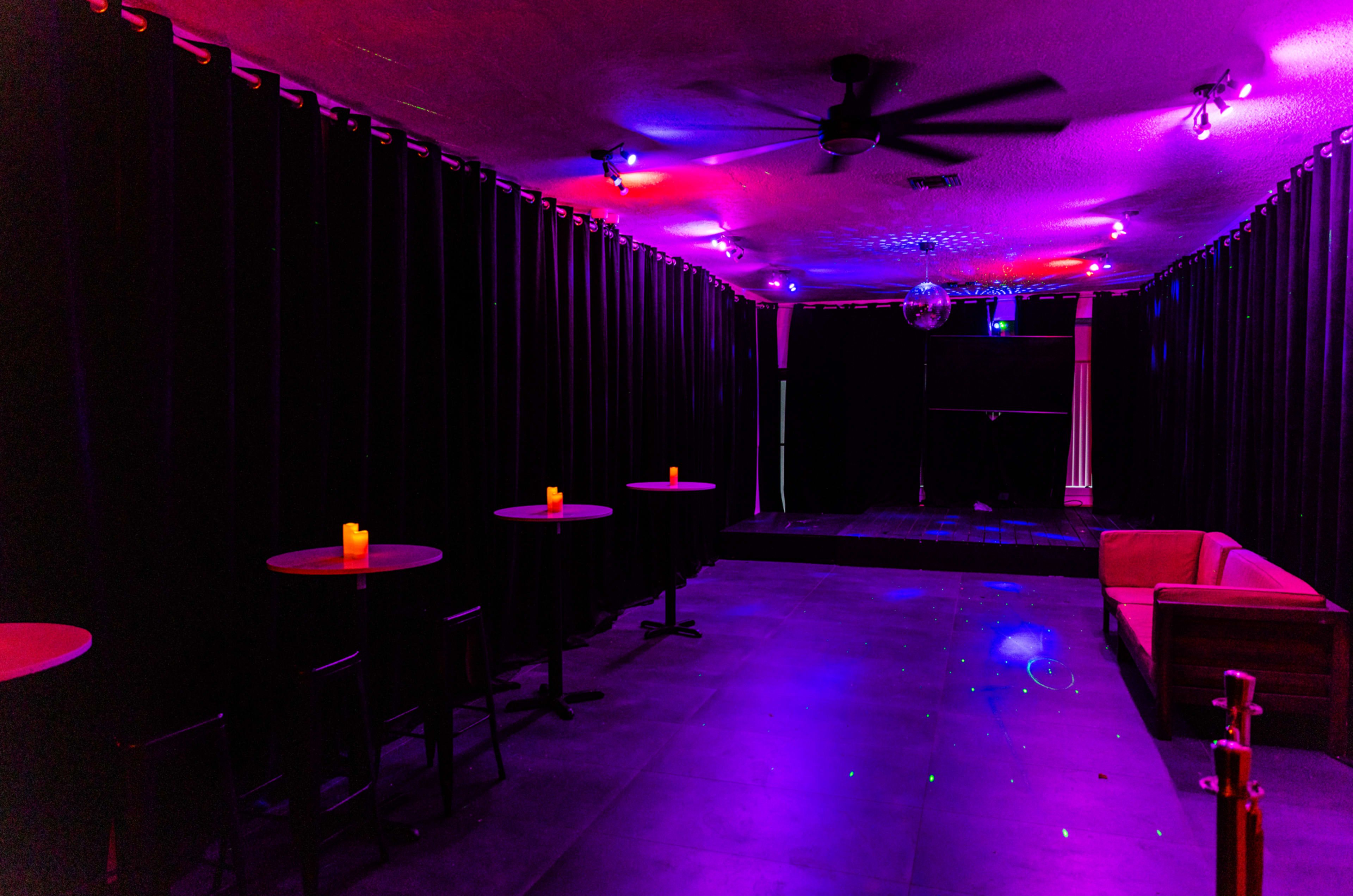 The image shows an empty room with dim lighting, featuring black curtains, small tables with candles, and a couch along one wall.