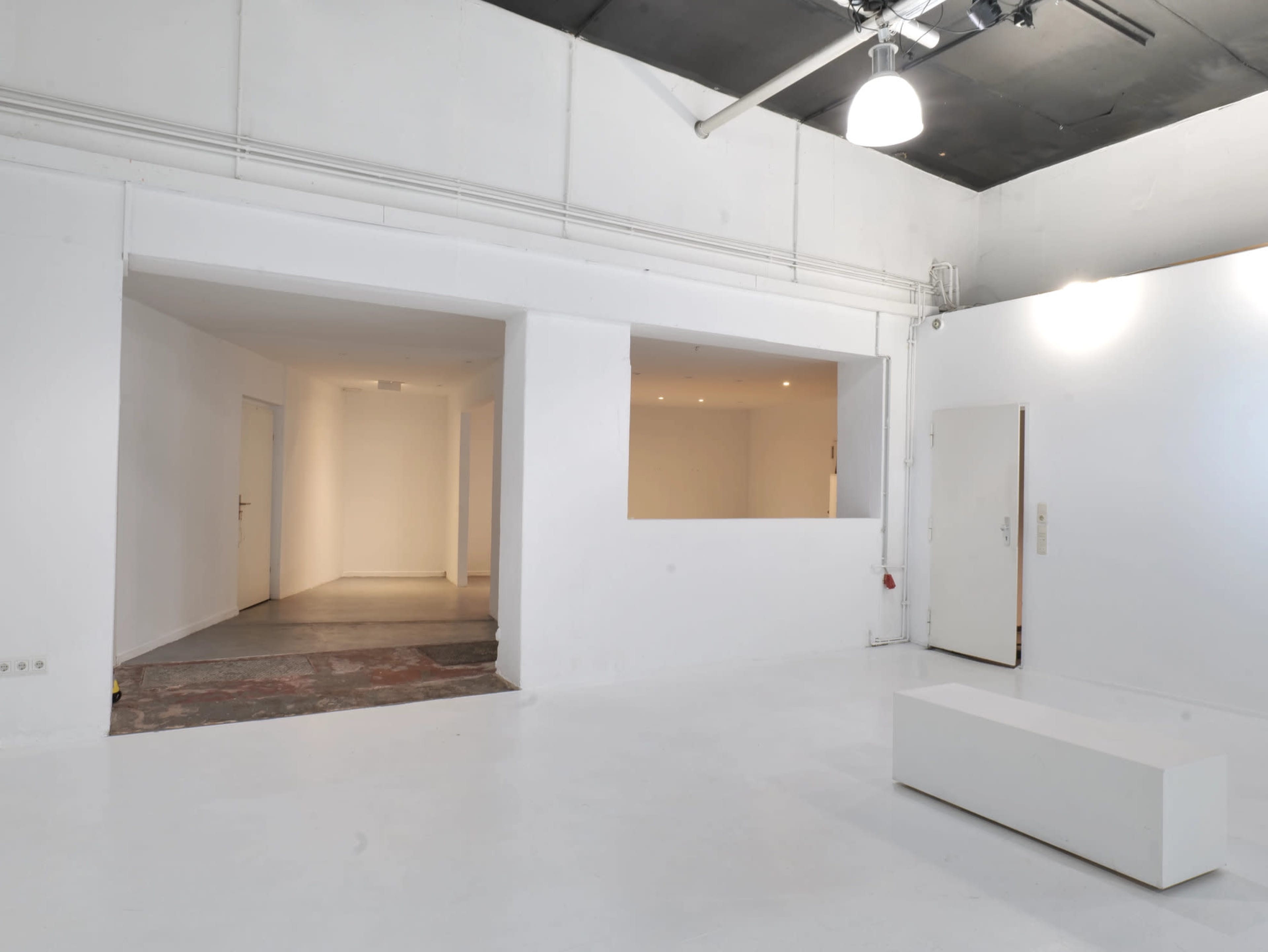 The image shows a bright, minimalist interior space with white walls, a light-colored floor, and an open area that leads to two doorways.