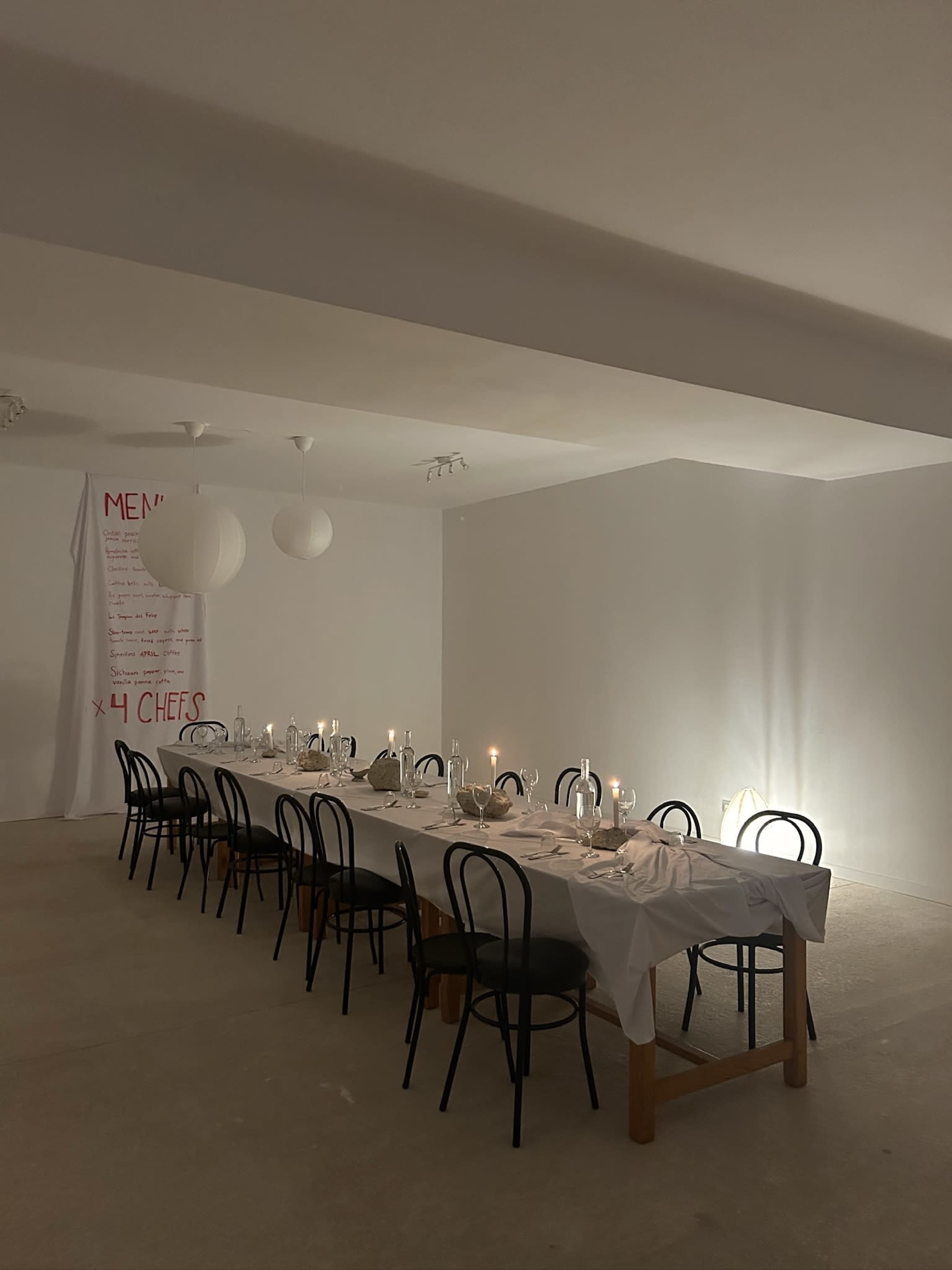 A long dining table set with plates and candles is positioned in a minimalist, softly lit room with white walls and contrasting black chairs.