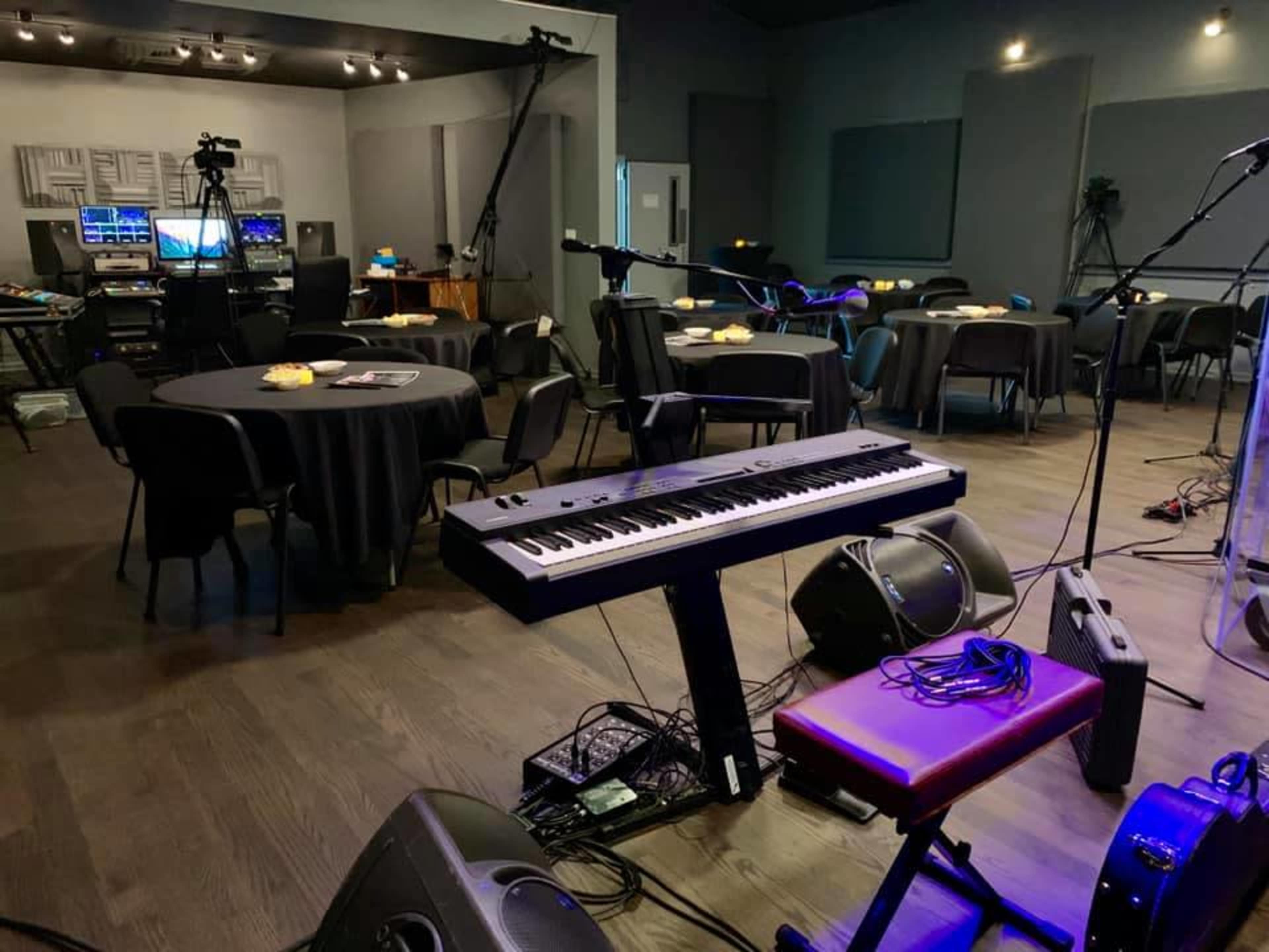 A music venue with a keyboard on a stand, surrounded by tables and chairs, and equipped with audio and video recording equipment.