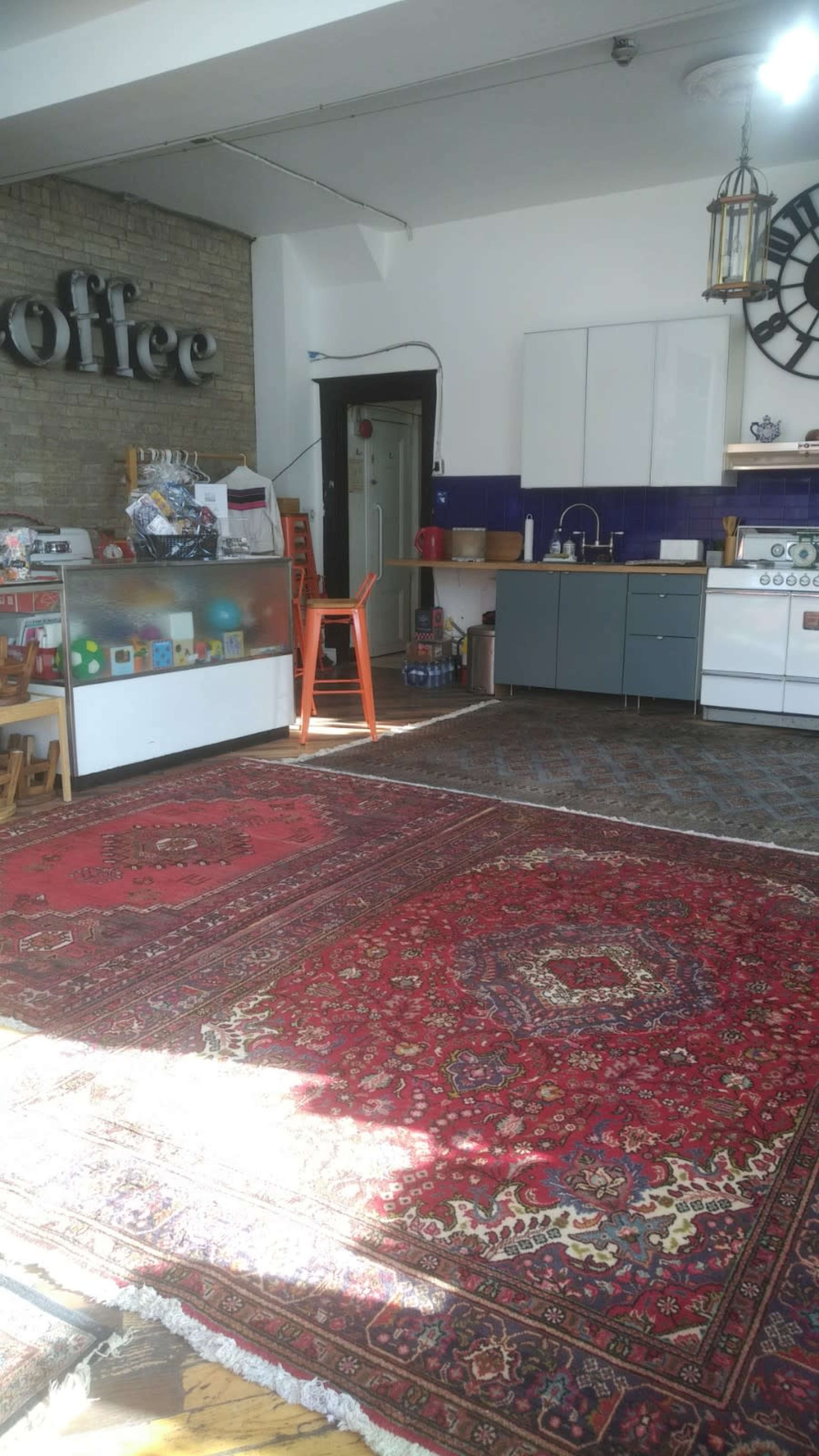 The image shows a spacious room with a coffee shop setup, featuring a counter with various items, a kitchen area, and large rugs on the wooden floor.