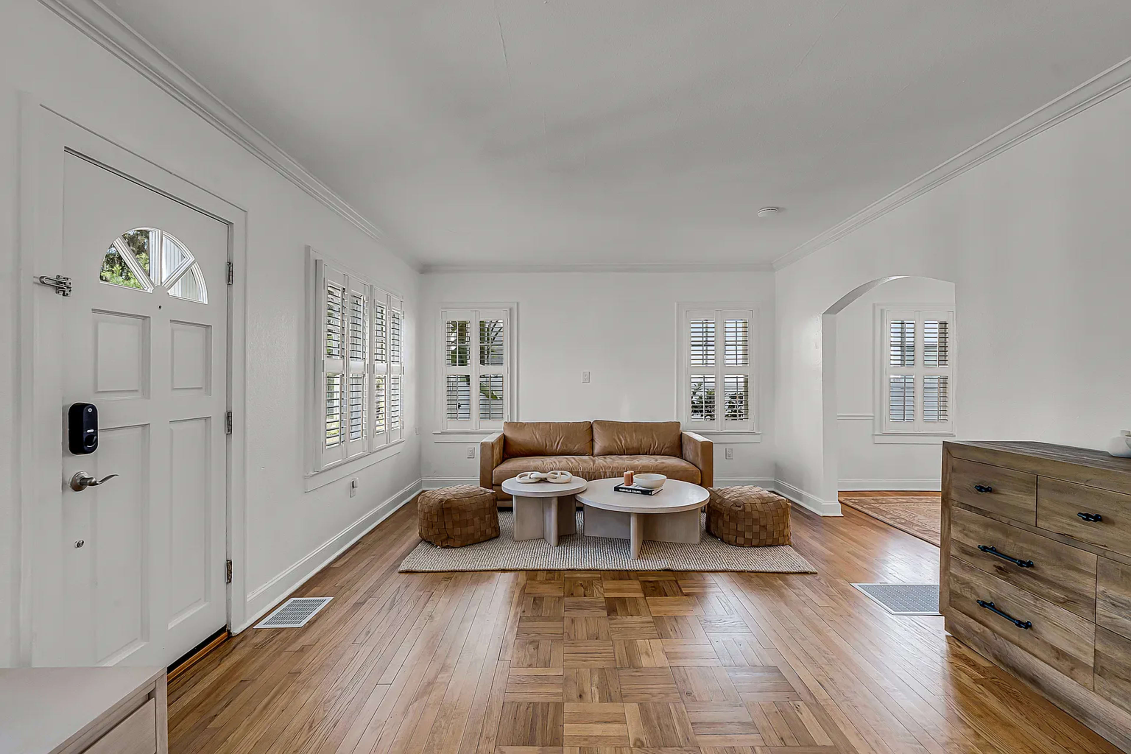 The image shows a bright, airy living room featuring light-colored walls, a brown leather couch, a round coffee table, and two poufs, with windows dressed in shutters.