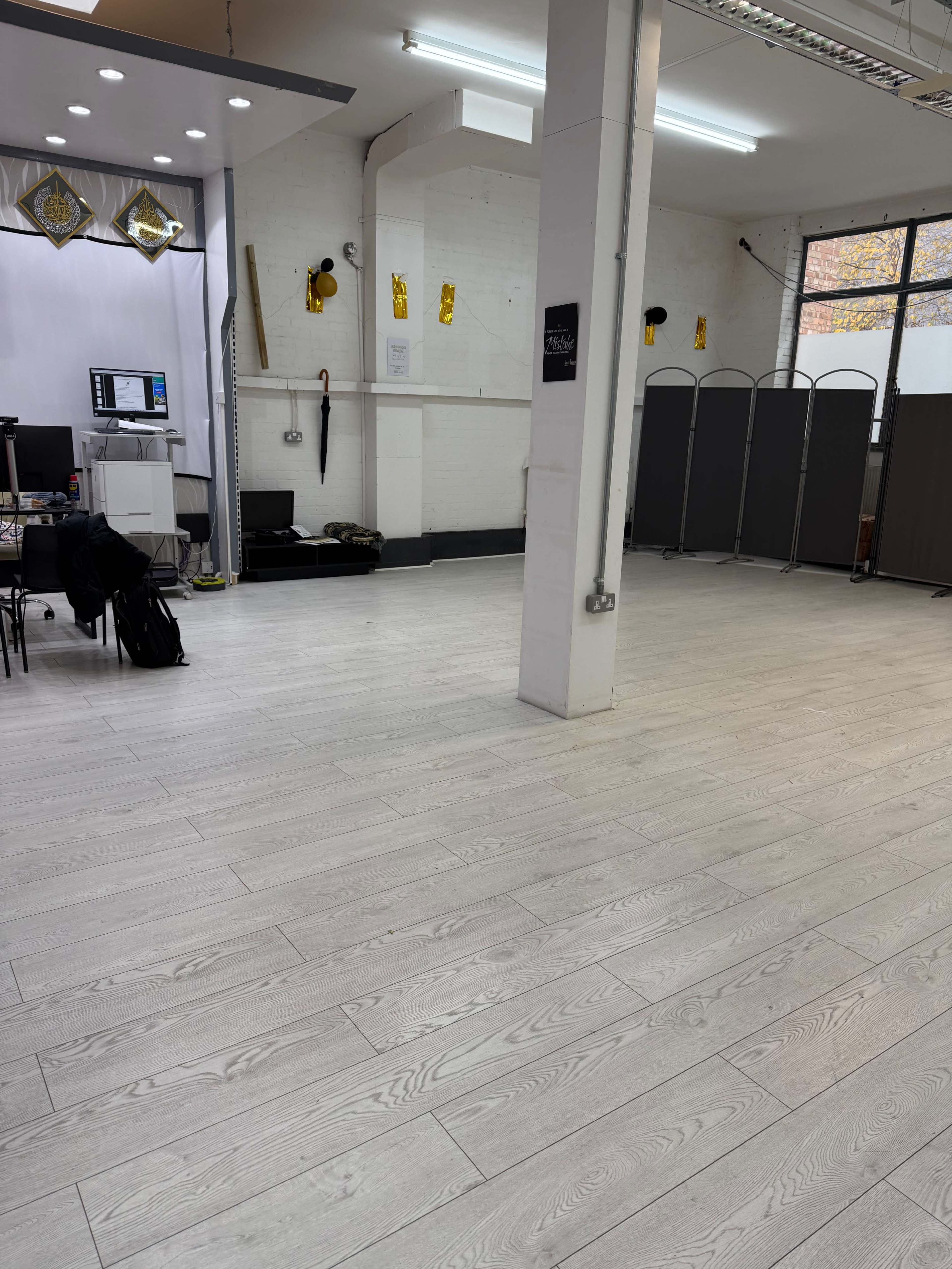 The image shows an empty indoor space with white wooden flooring, a partitioned area in the background, and a desk setup with a computer on one side.
