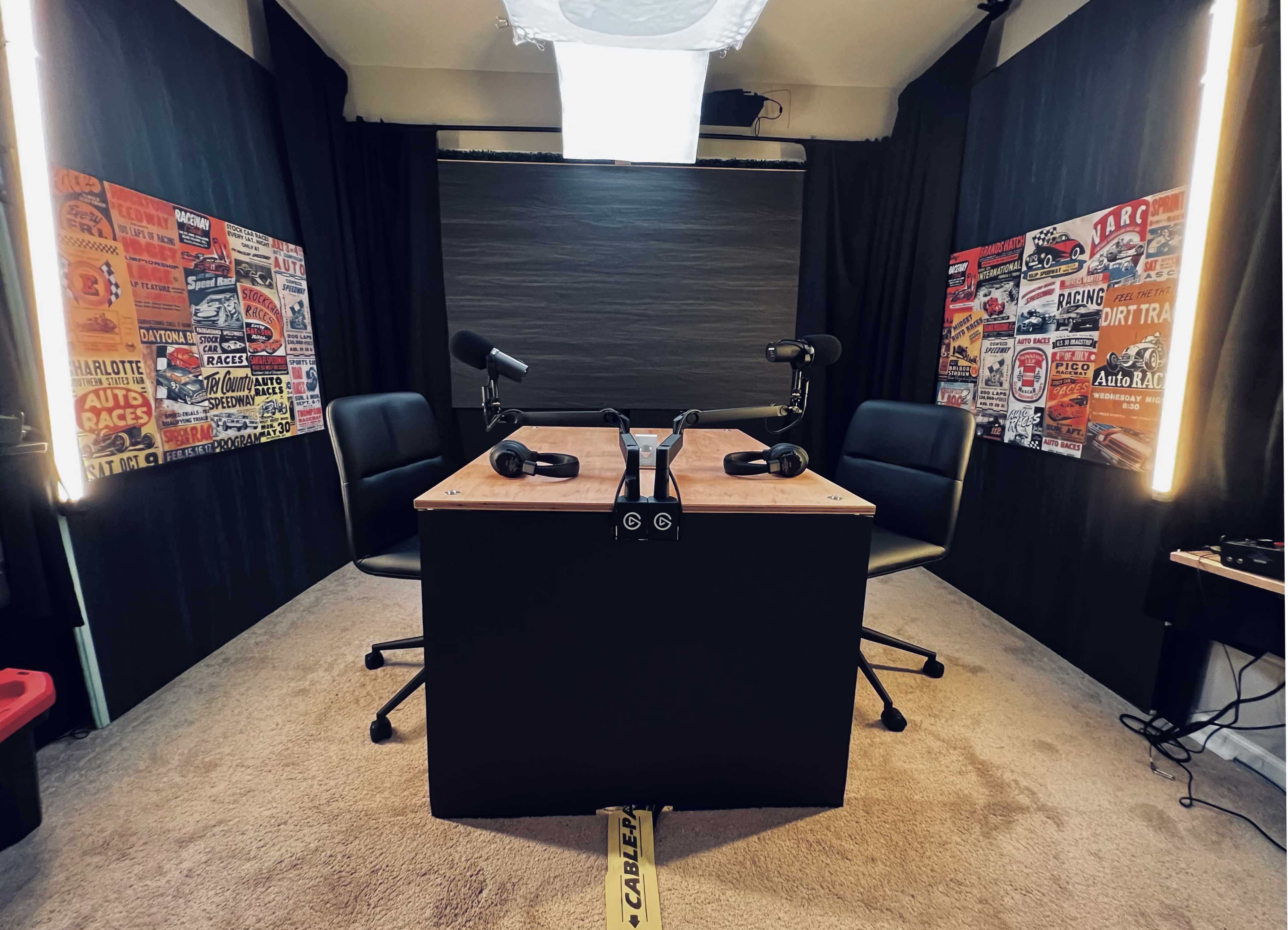 A podcast recording studio featuring a table with two chairs, microphones, and walls adorned with vintage racing posters.
