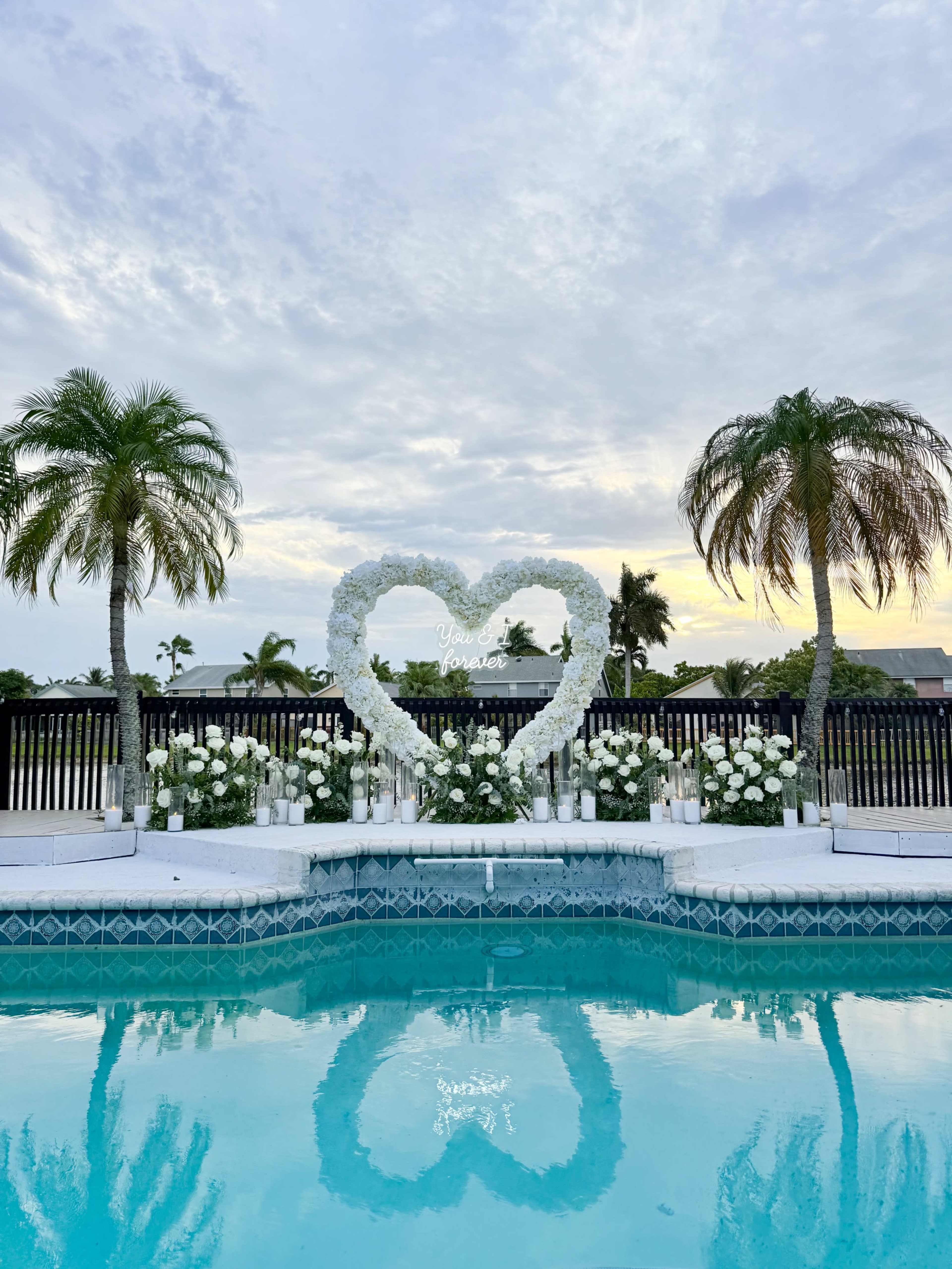 A heart-shaped flower arrangement stands beside a pool, framed by palm trees and surrounded by white floral decorations.