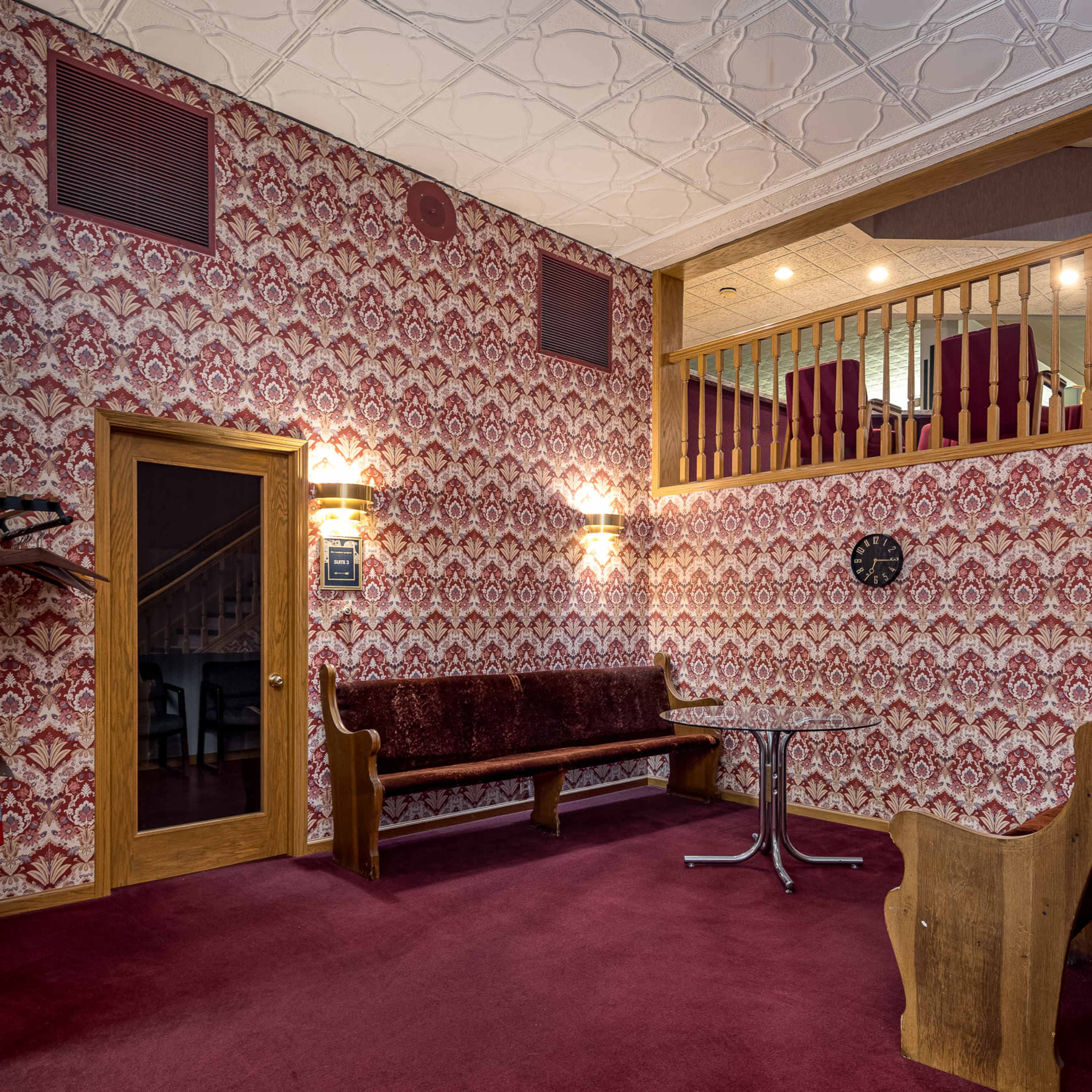 A room with patterned wallpaper, a red carpet, a wooden entrance door, a round table, and a seating area along one wall.