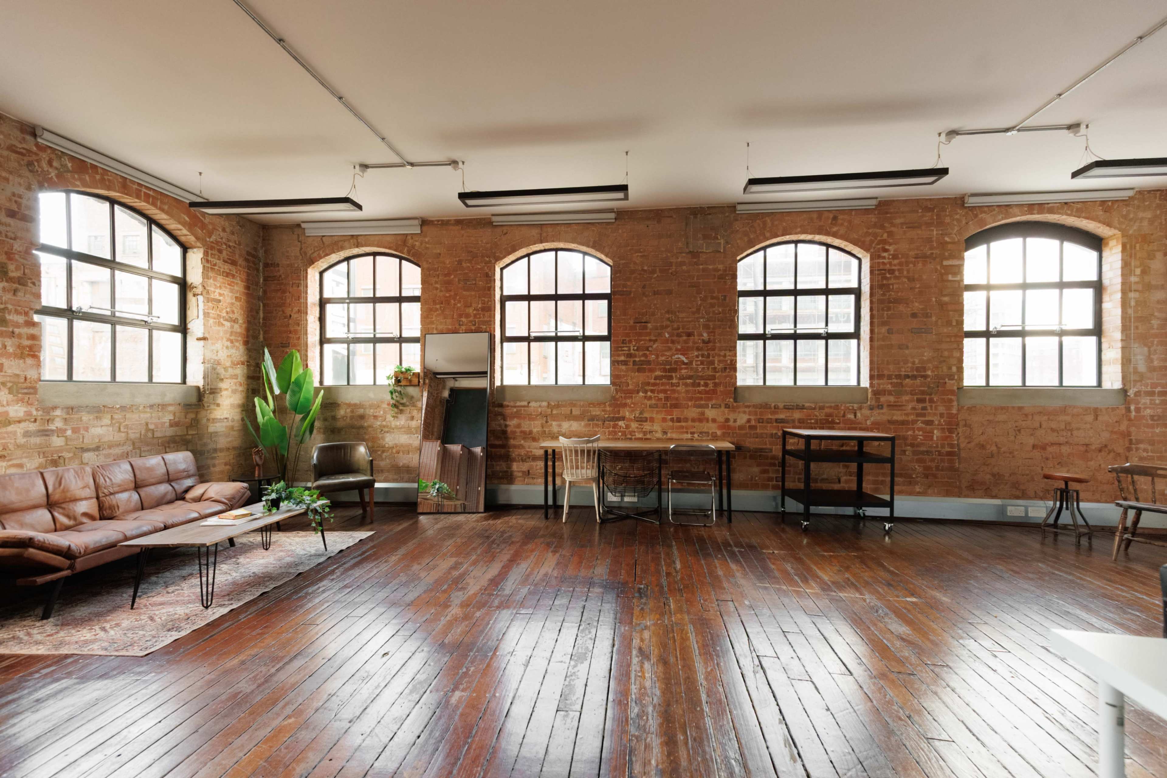 The image shows a spacious interior featuring exposed brick walls, large windows, wooden floors, a leather sofa, a dining table, and various plants.