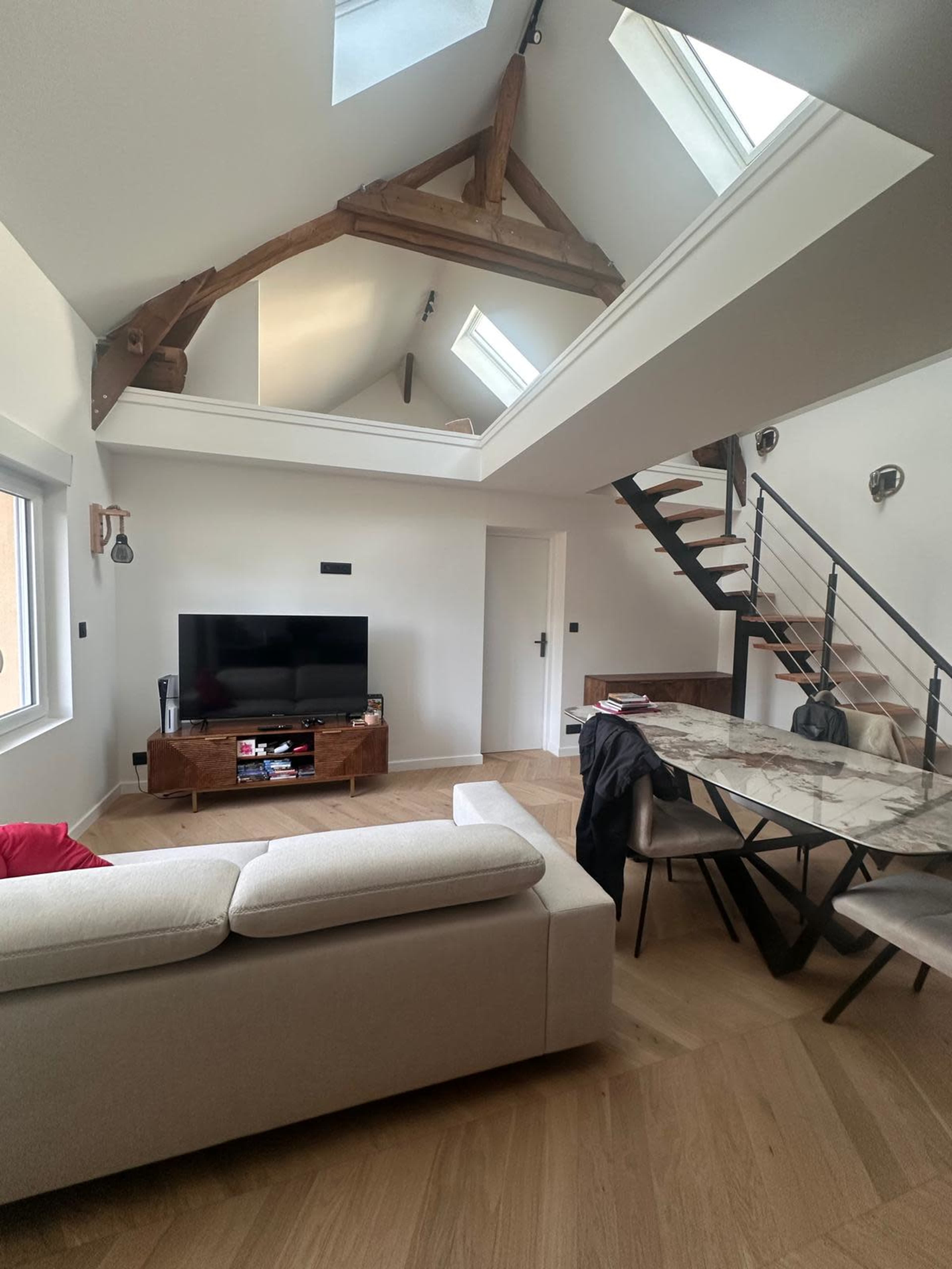 A modern living room features a sectional sofa, a wooden television stand, a dining table, and a staircase leading to an upper level, all under a sloped ceiling with skylights.