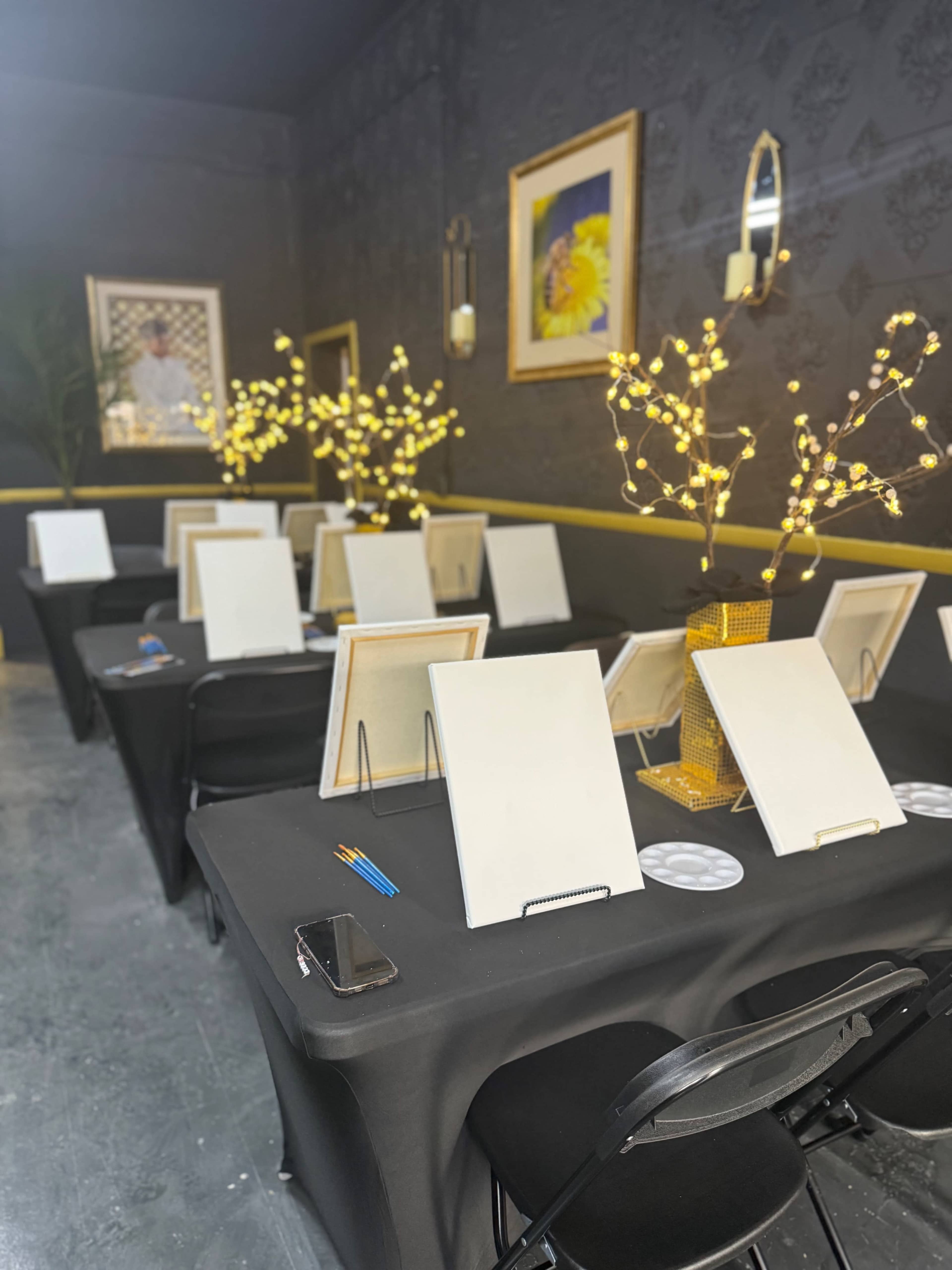 A series of tables covered with black cloth are set up for a painting event, each featuring a blank canvas and artistic supplies against a decorated backdrop.