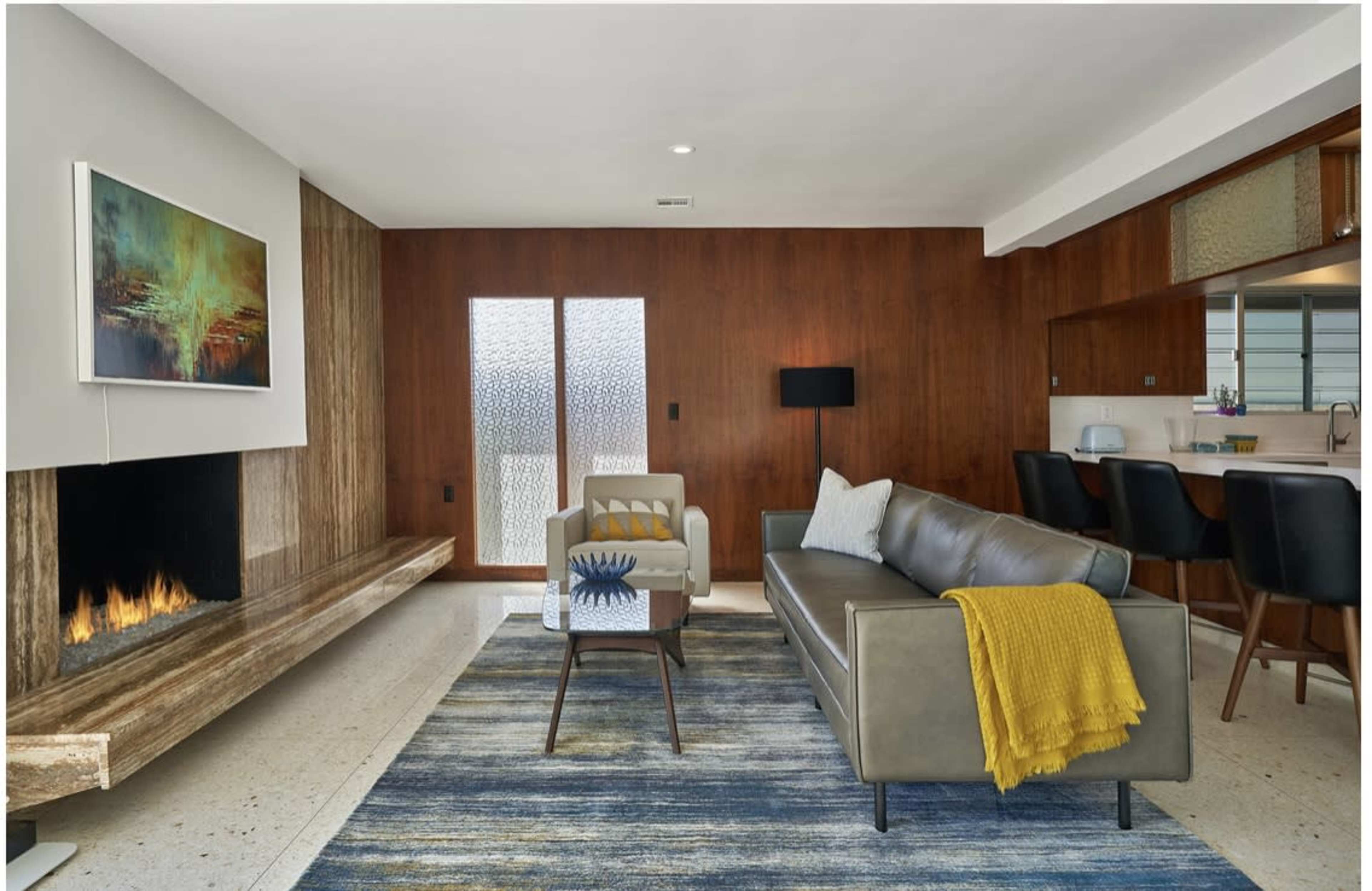 A modern living room with a fireplace, a gray sofa, a white armchair, and a blue and yellow rug, set against wooden wall panels and a kitchen area in the background.