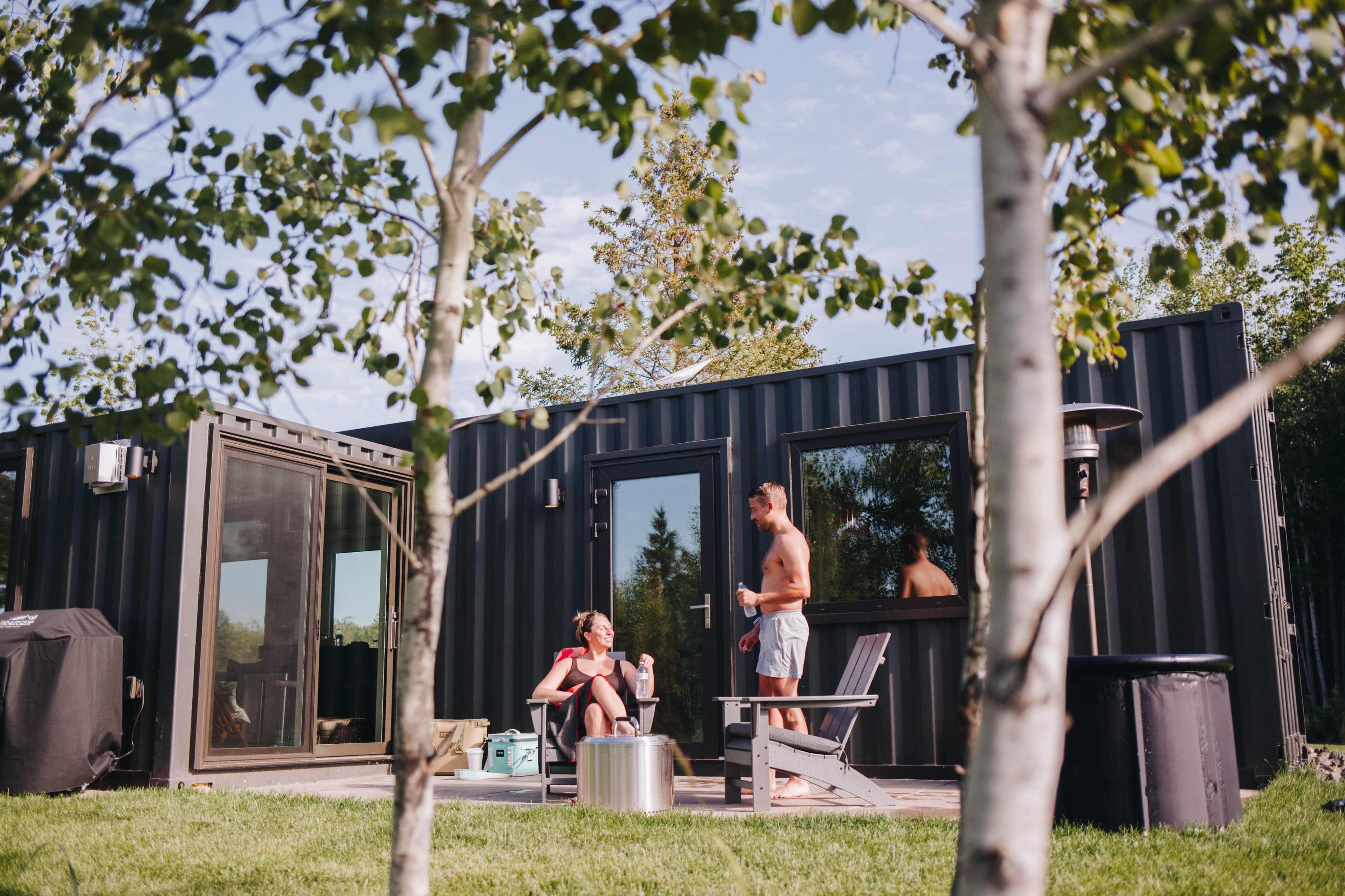 Two people converse outside a modern container home, with one seated and the other standing near the entrance.