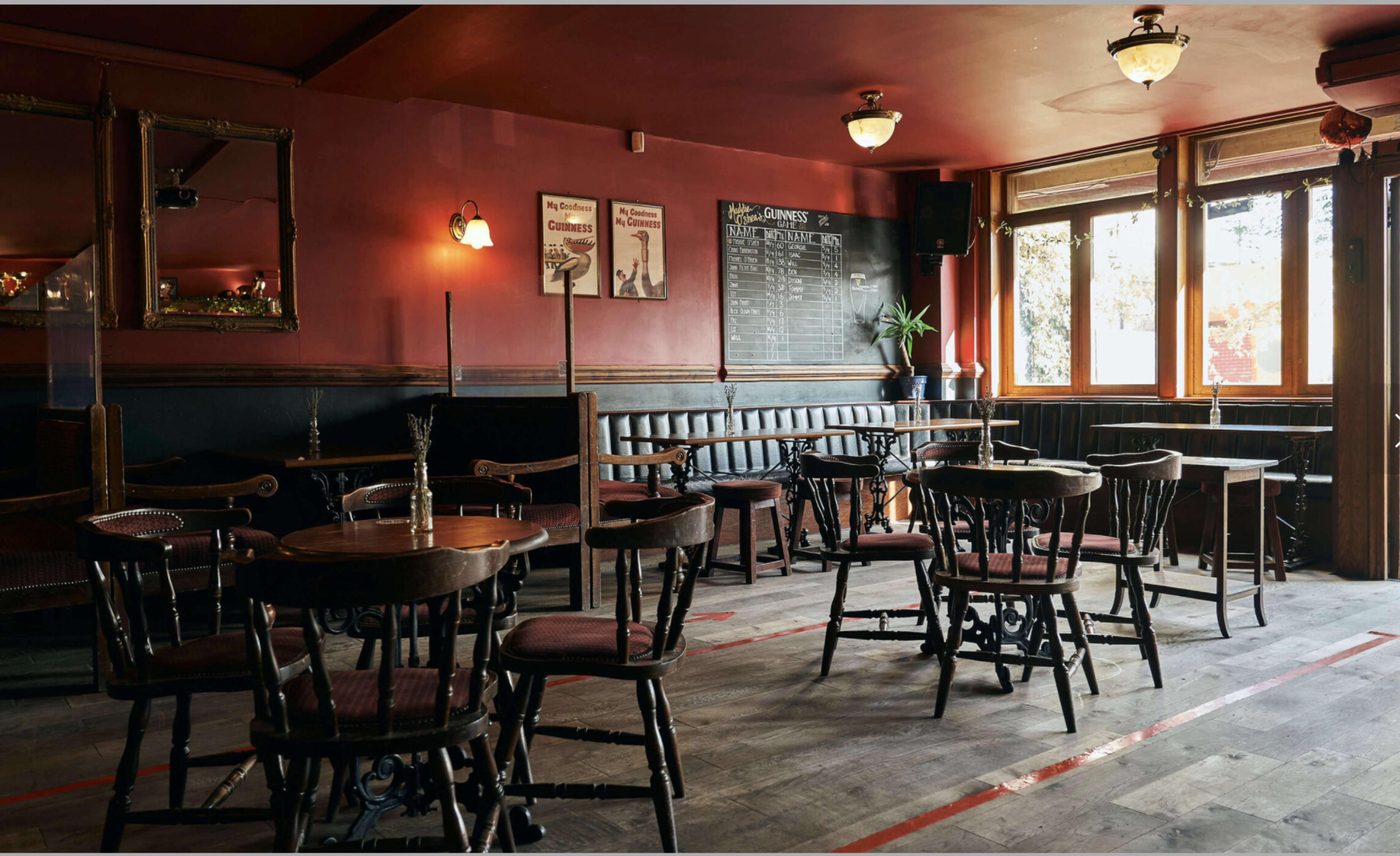 A vintage-style pub interior with wooden tables and chairs arranged around the space, decorative mirrors on the walls, and a chalkboard menu near the large windows.