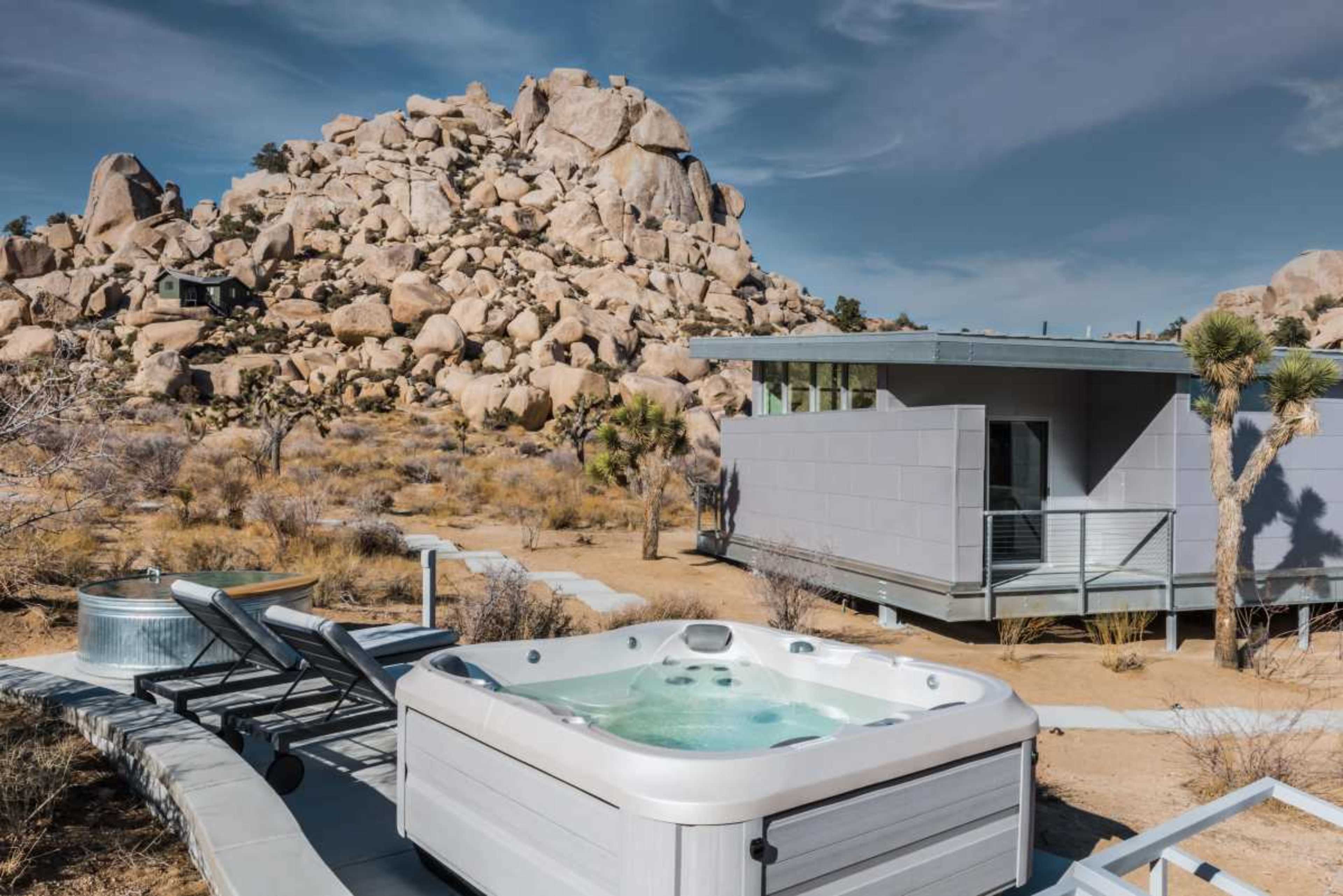 A hot tub sits in front of a modern gray building, surrounded by rocky terrain and sparse vegetation.