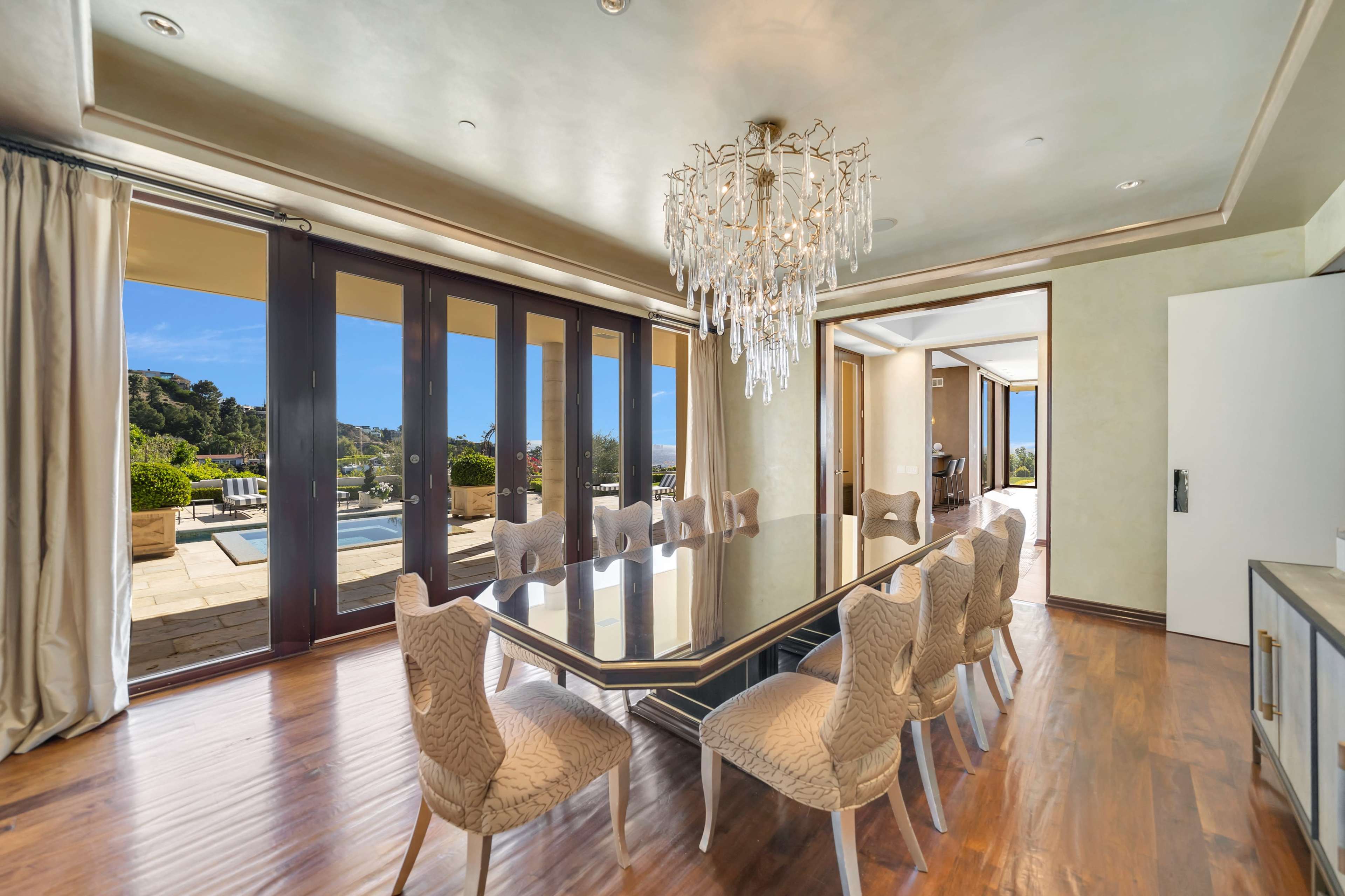 A modern dining room features a large glass table surrounded by elegantly patterned chairs, with a chandelier above and glass doors opening to a patio and pool area.