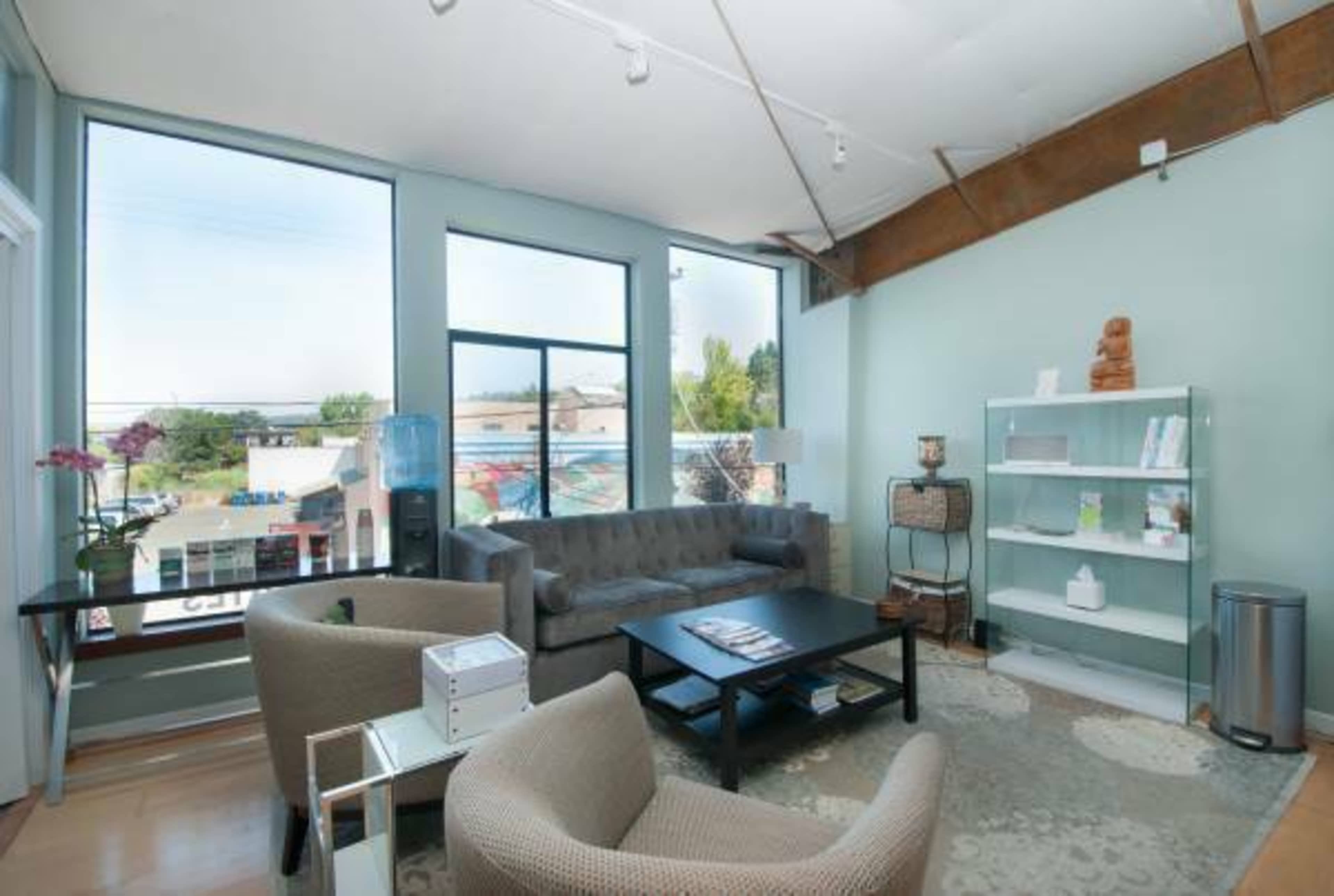 A well-lit, modern lounge area with large windows, a seating arrangement that includes two chairs and a sofa, a coffee table, and a shelf with various items.