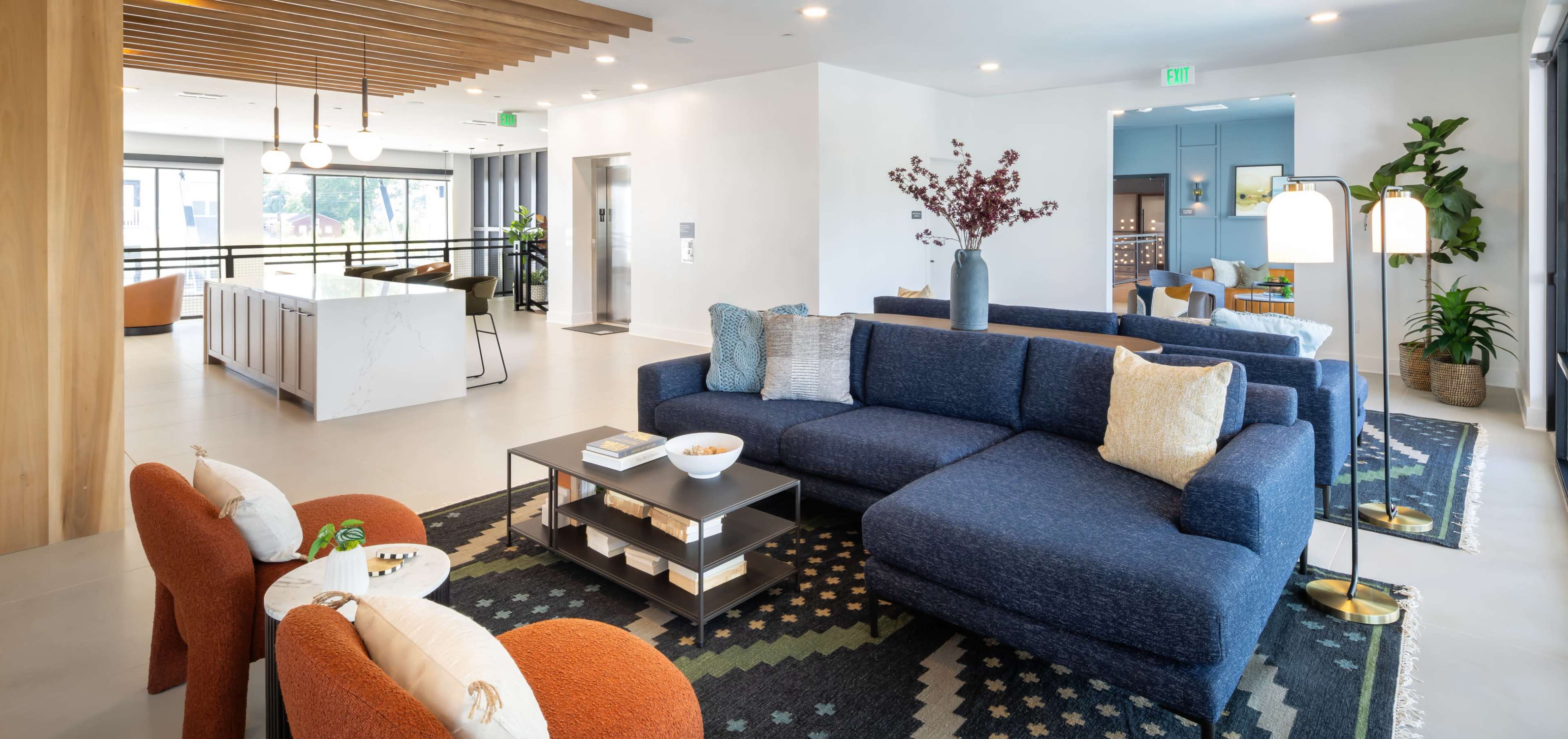 The image depicts a modern, open living space featuring a blue sectional sofa, two orange armchairs, and a kitchen area in the background, along with plants and decorative elements.