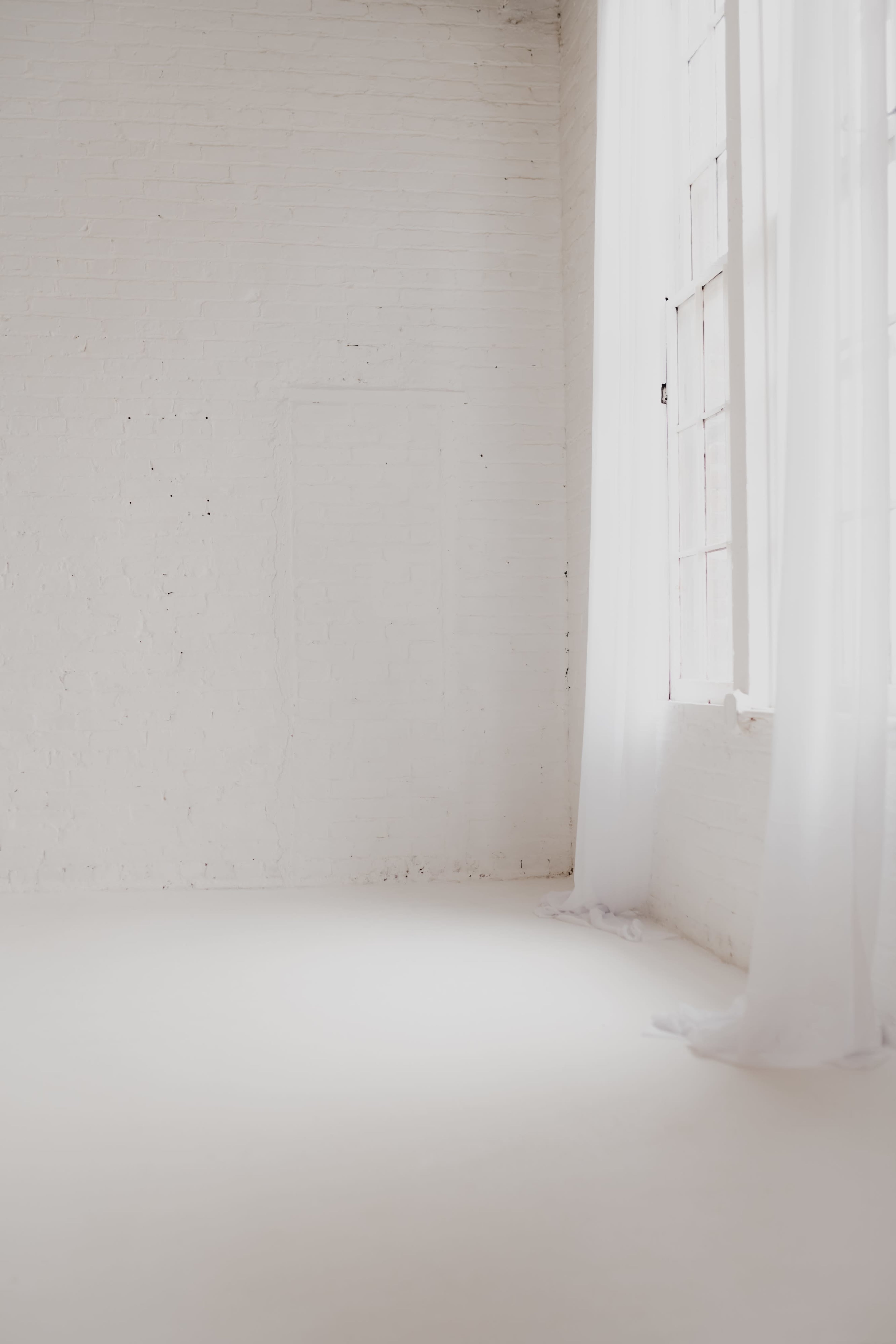 The image shows a bright, empty room with white brick walls and sheer curtains near a window.