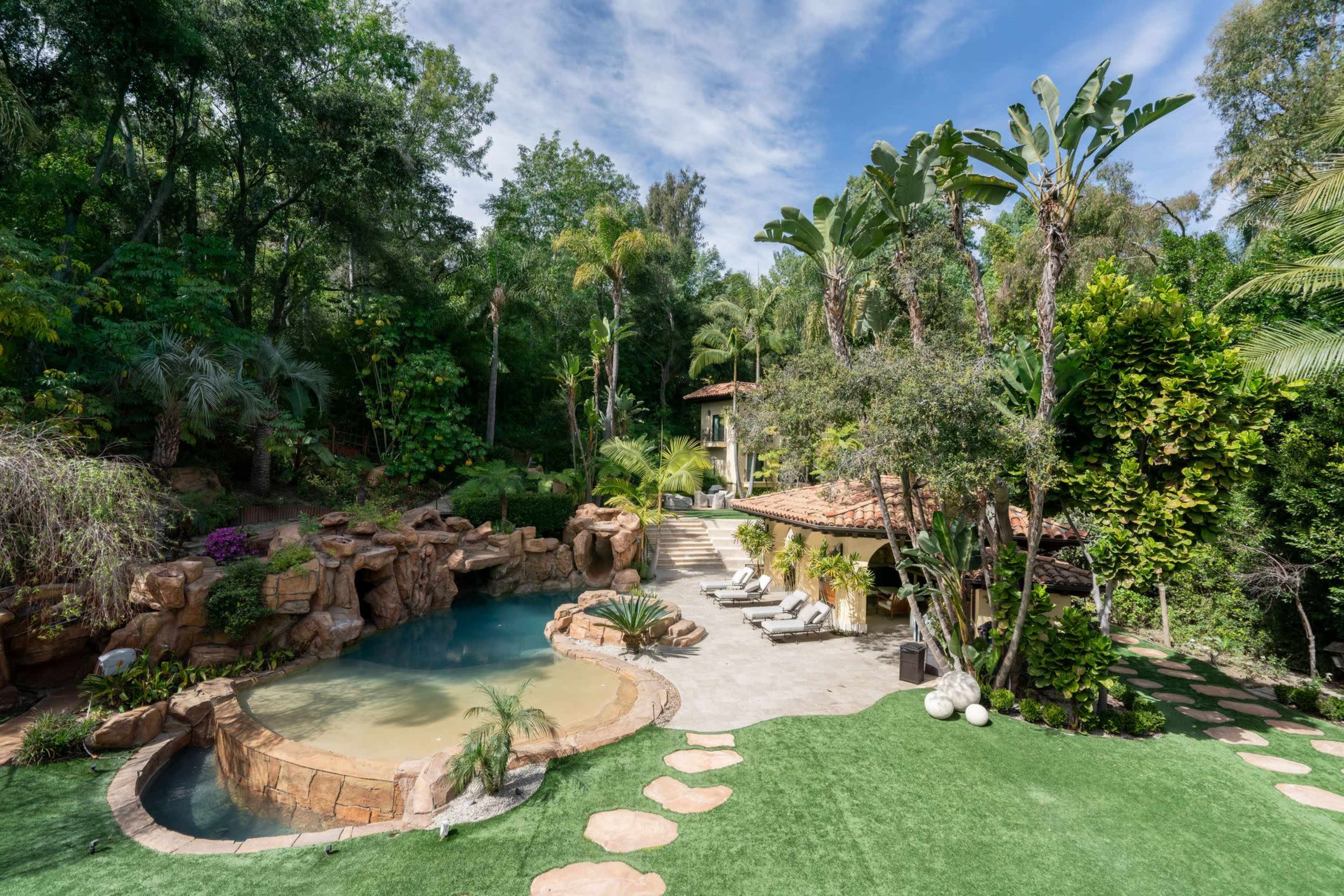 The image shows a private outdoor space featuring a landscaped pool area surrounded by lush vegetation, palm trees, and a stone pathway.