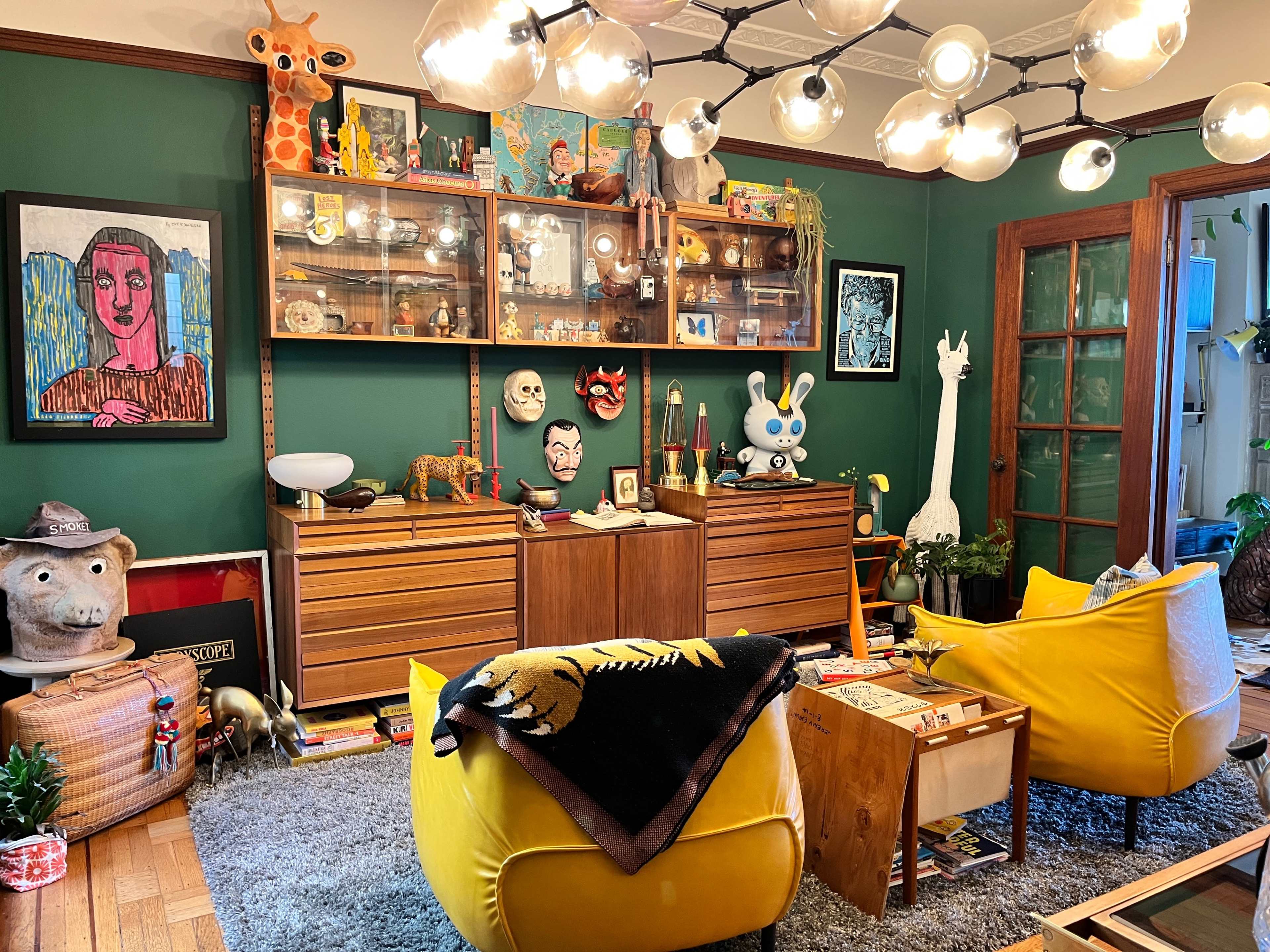 A colorful living room with yellow armchairs, a green wall, and a wooden cabinet displaying various art pieces and collectibles.