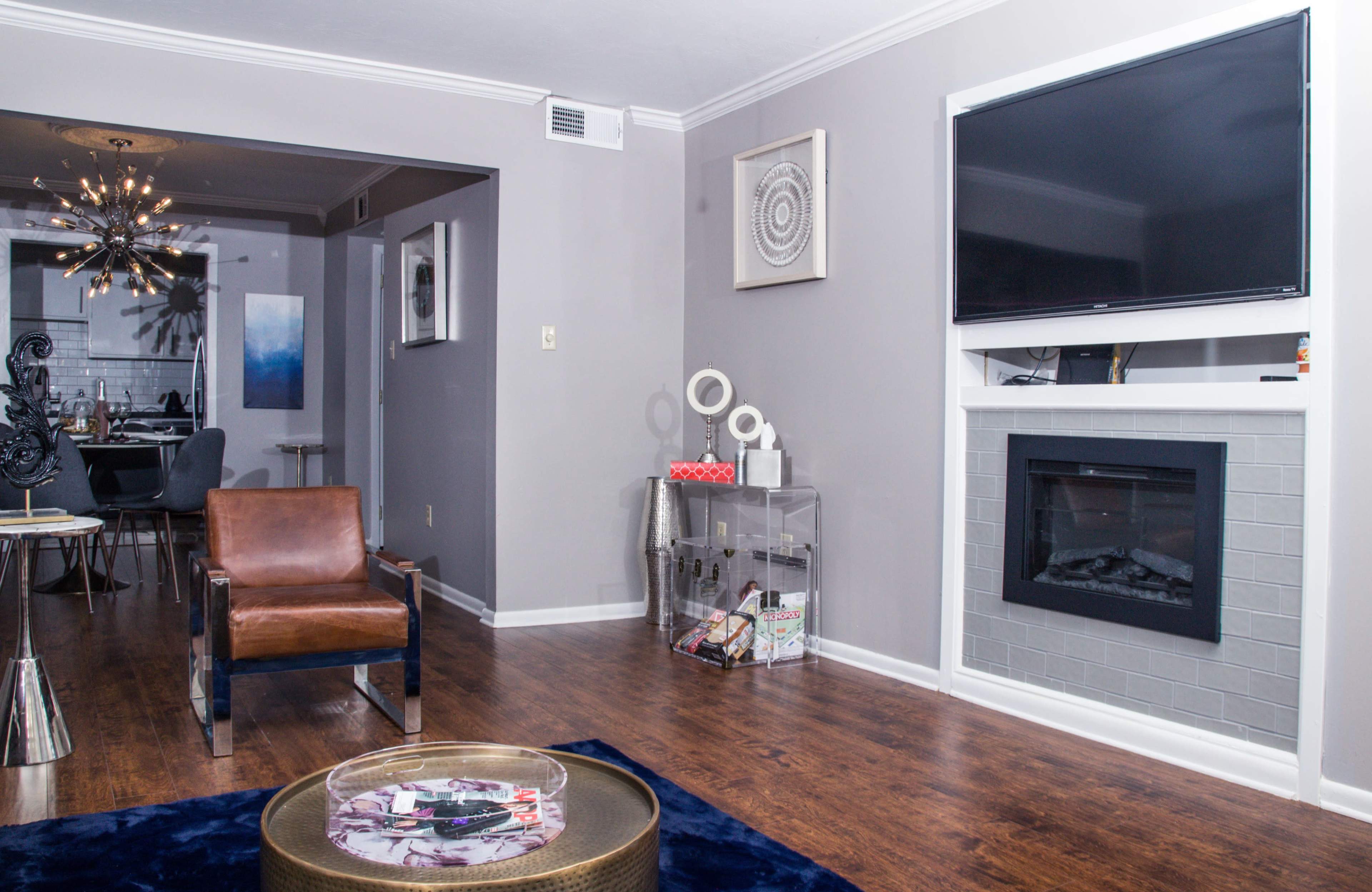 A modern living room features a brown armchair, a gray fireplace with a television mounted above, and a coffee table on a blue rug, with a dining area visible in the background.