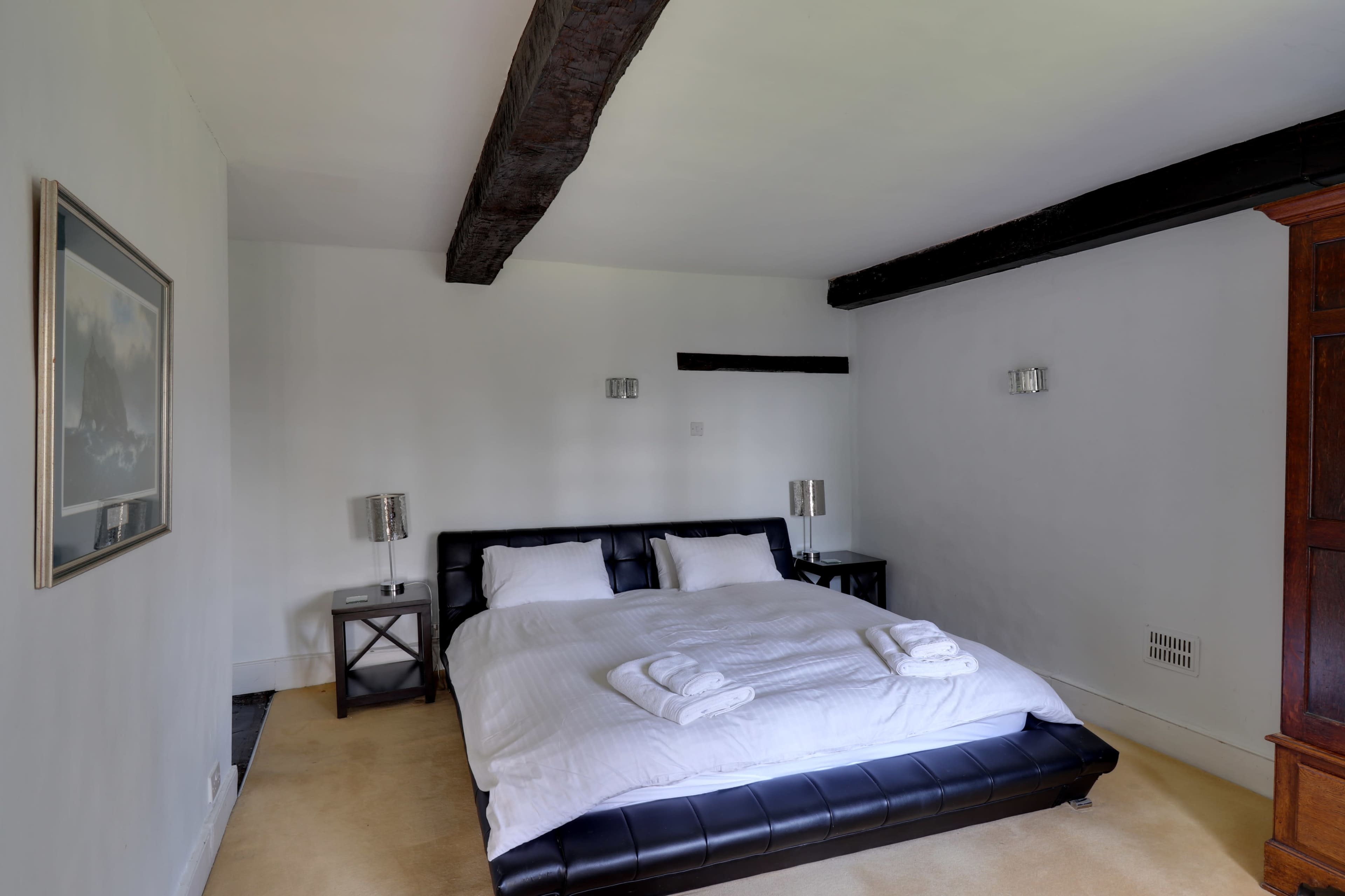 A spacious bedroom features a black leather bed with white linens and two bedside lamps.