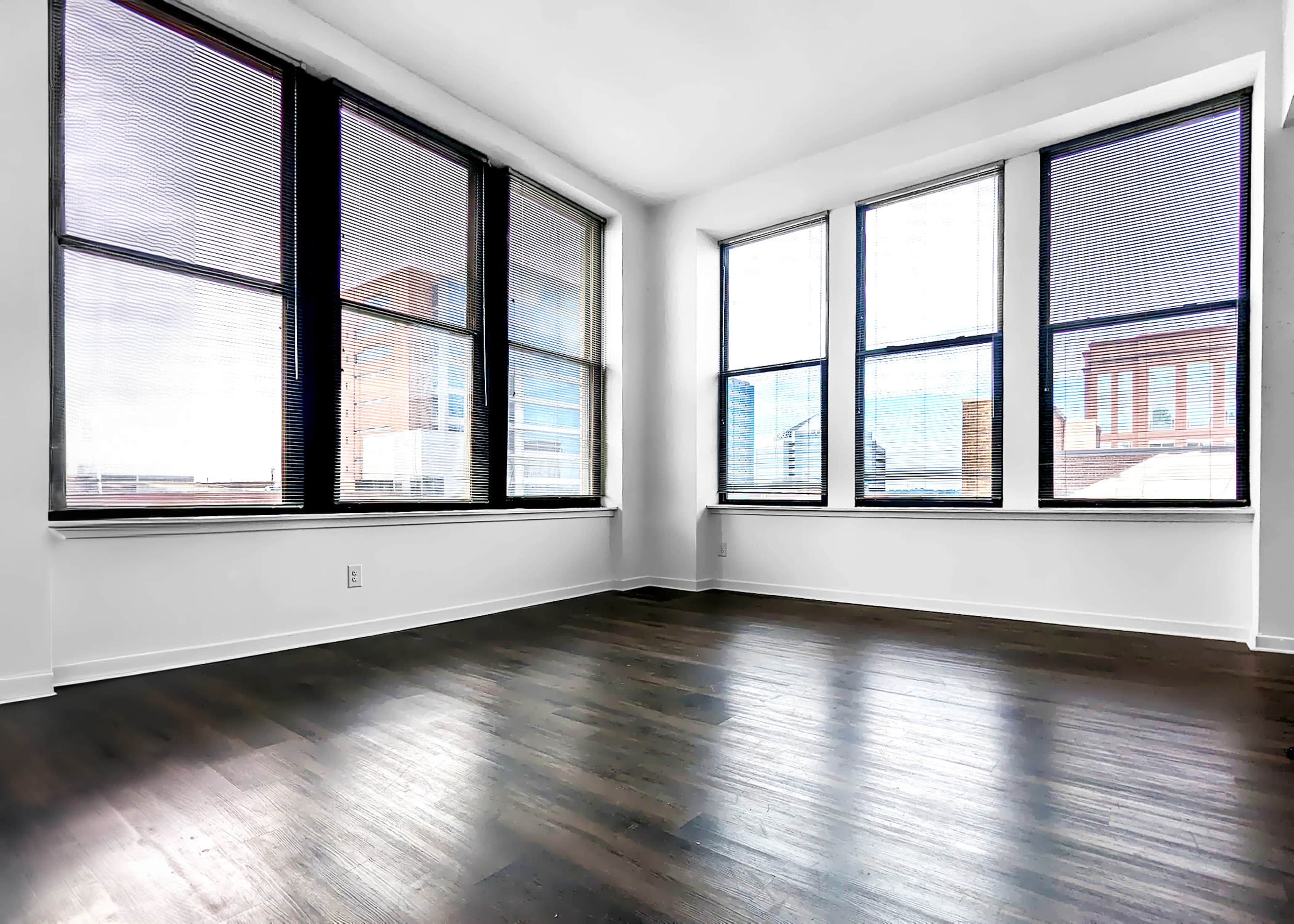 A spacious room with large windows, showcasing city views and a polished wooden floor.
