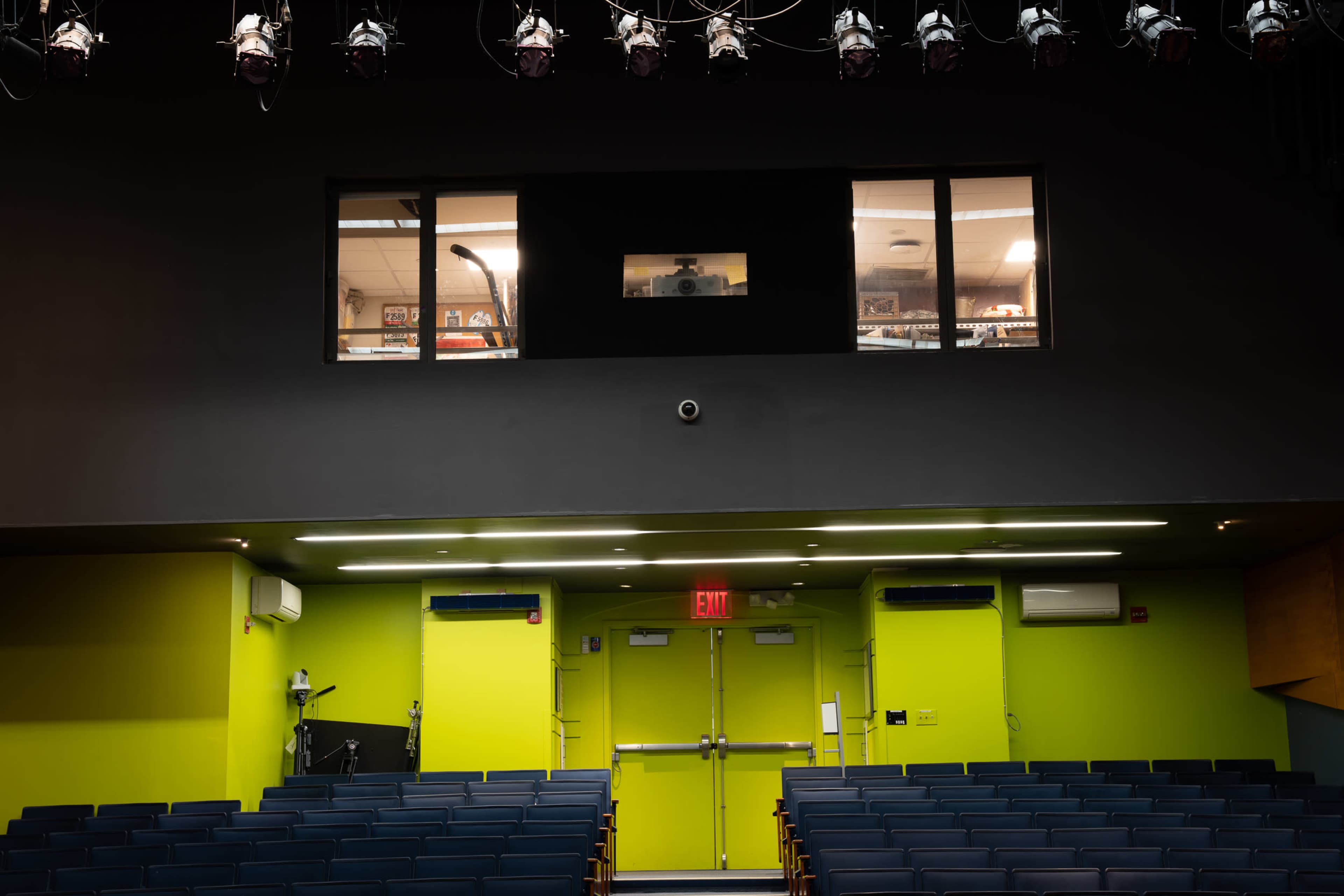 An empty auditorium with rows of blue seats and a green-lit stage area, featuring a control room window and emergency exit doors.