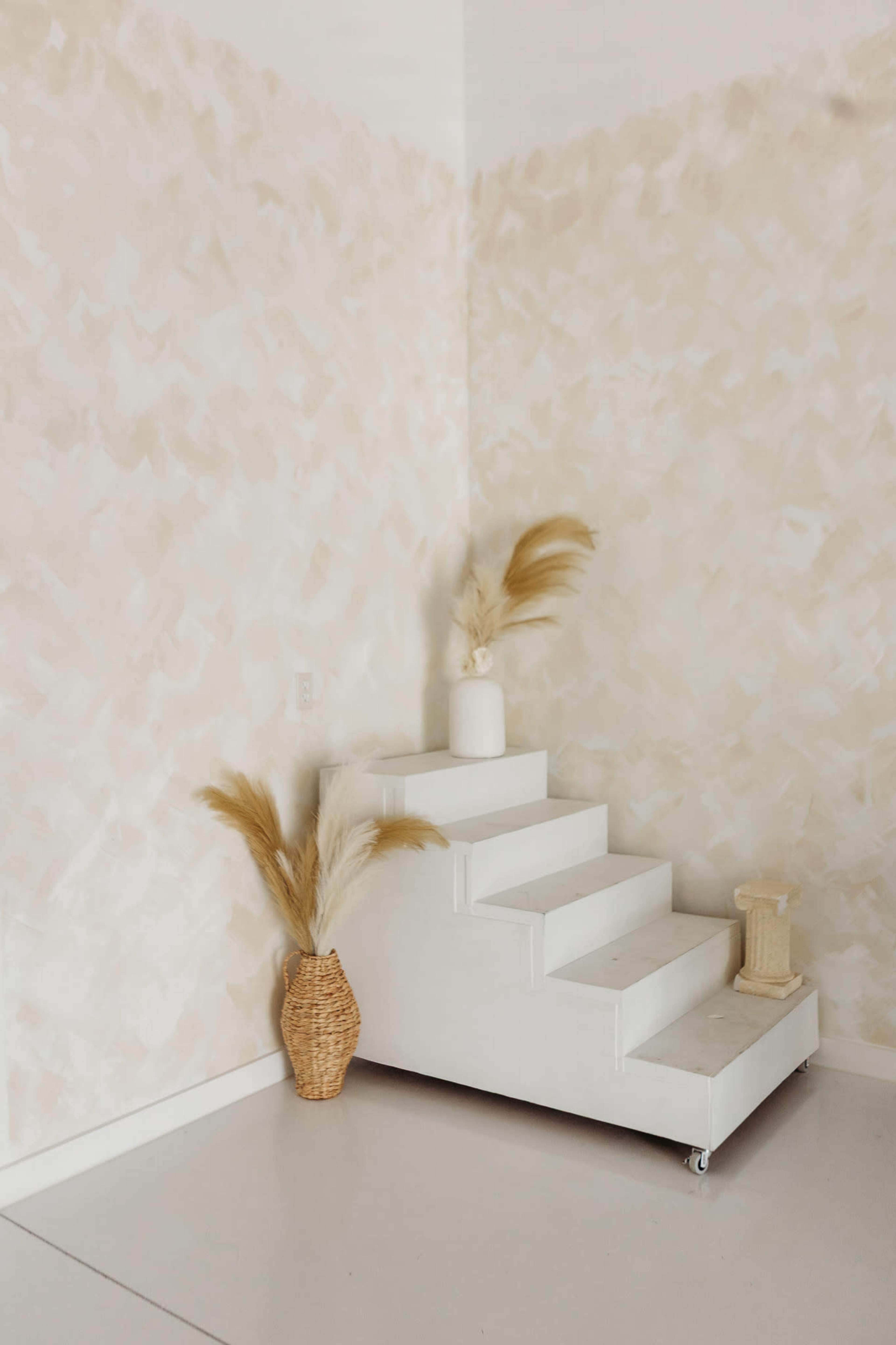 A minimalist corner features a white staircase on wheels beside a woven basket and a vase with dried pampas grass against a textured, light-colored wall.