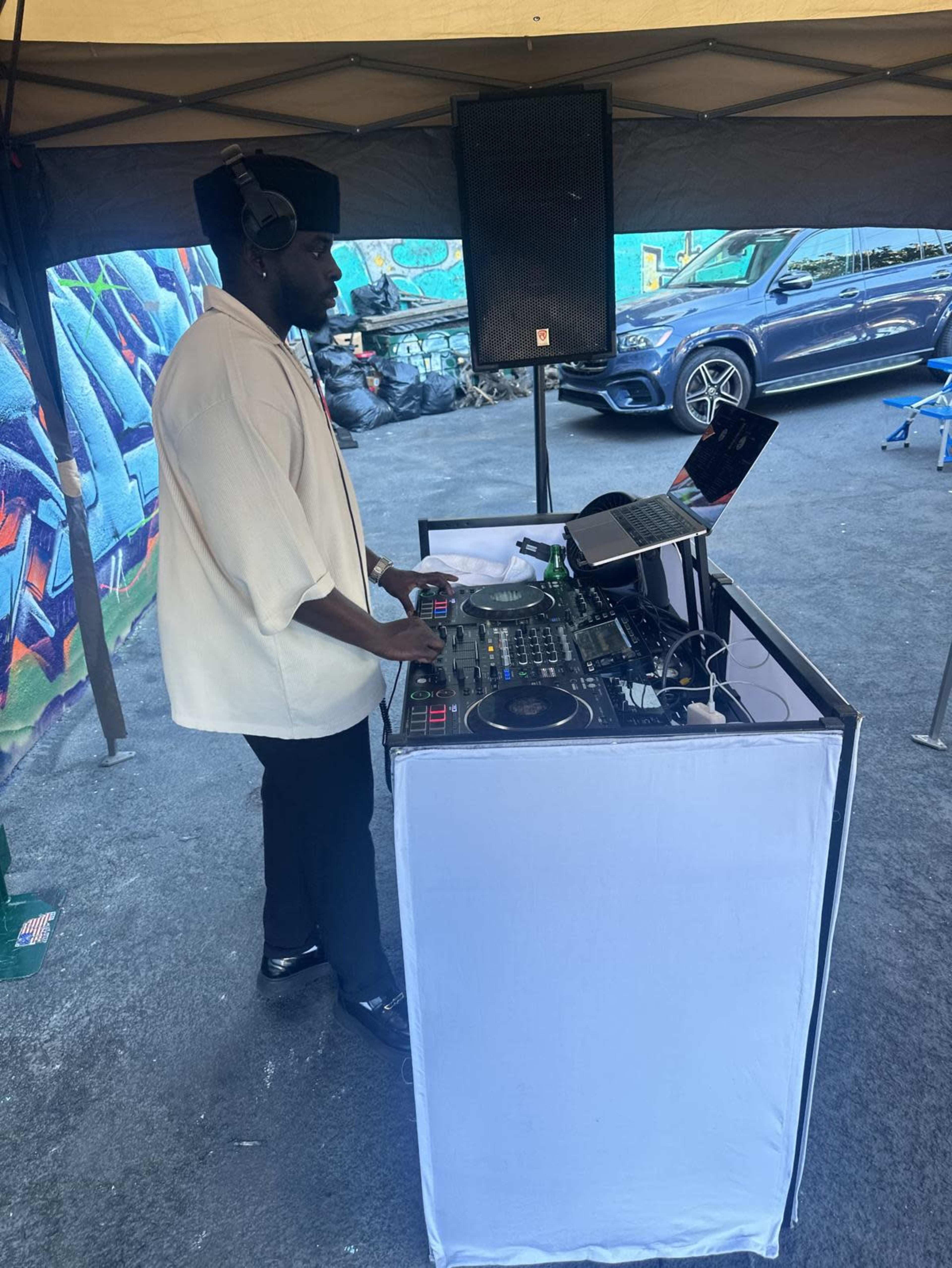 A DJ stands behind a table with turntables and a laptop, performing at an outdoor event under a canopy.
