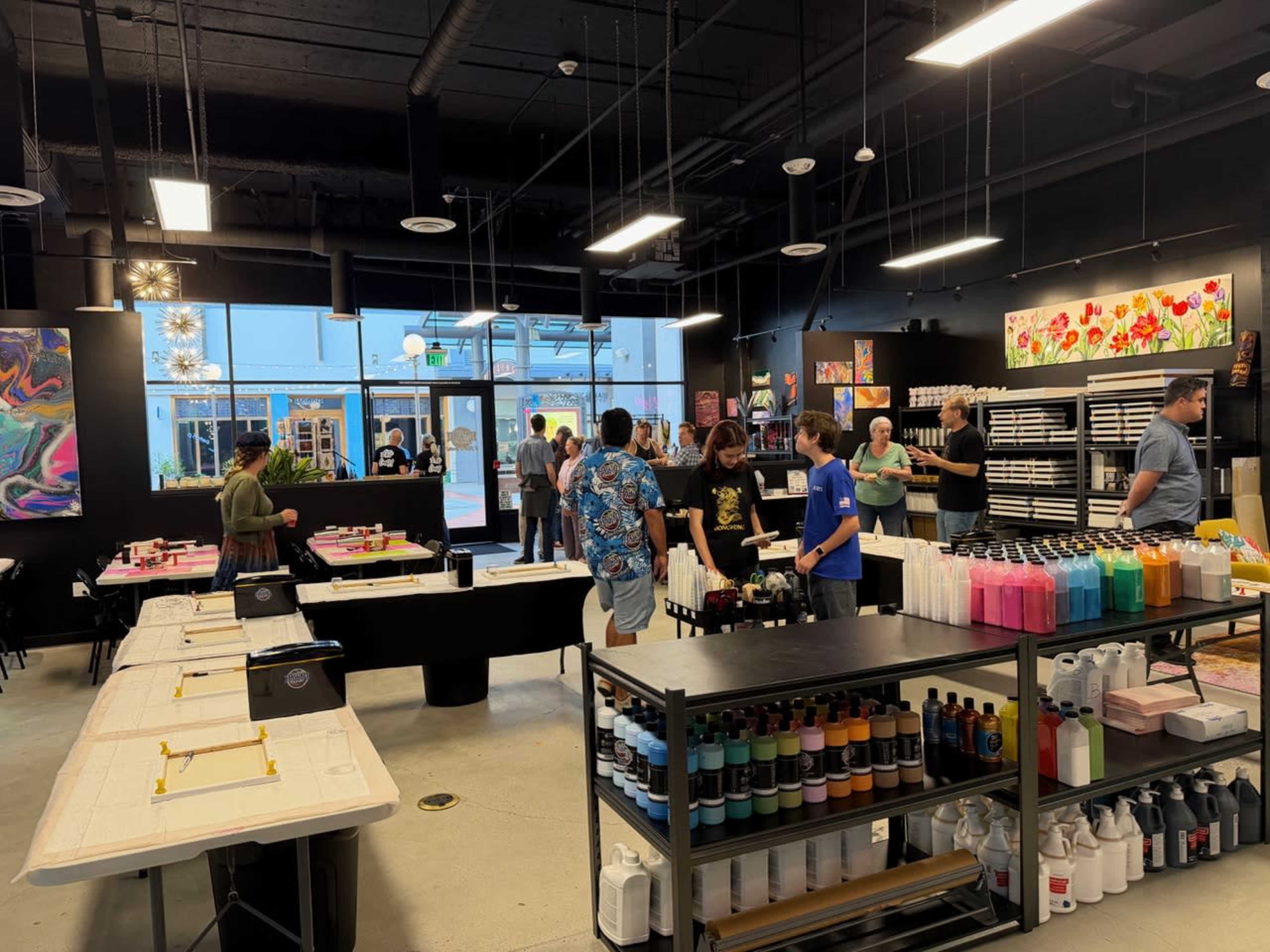 The image depicts a vibrant art studio with several people engaged in creative activities, surrounded by shelves stocked with colorful paints and art supplies.