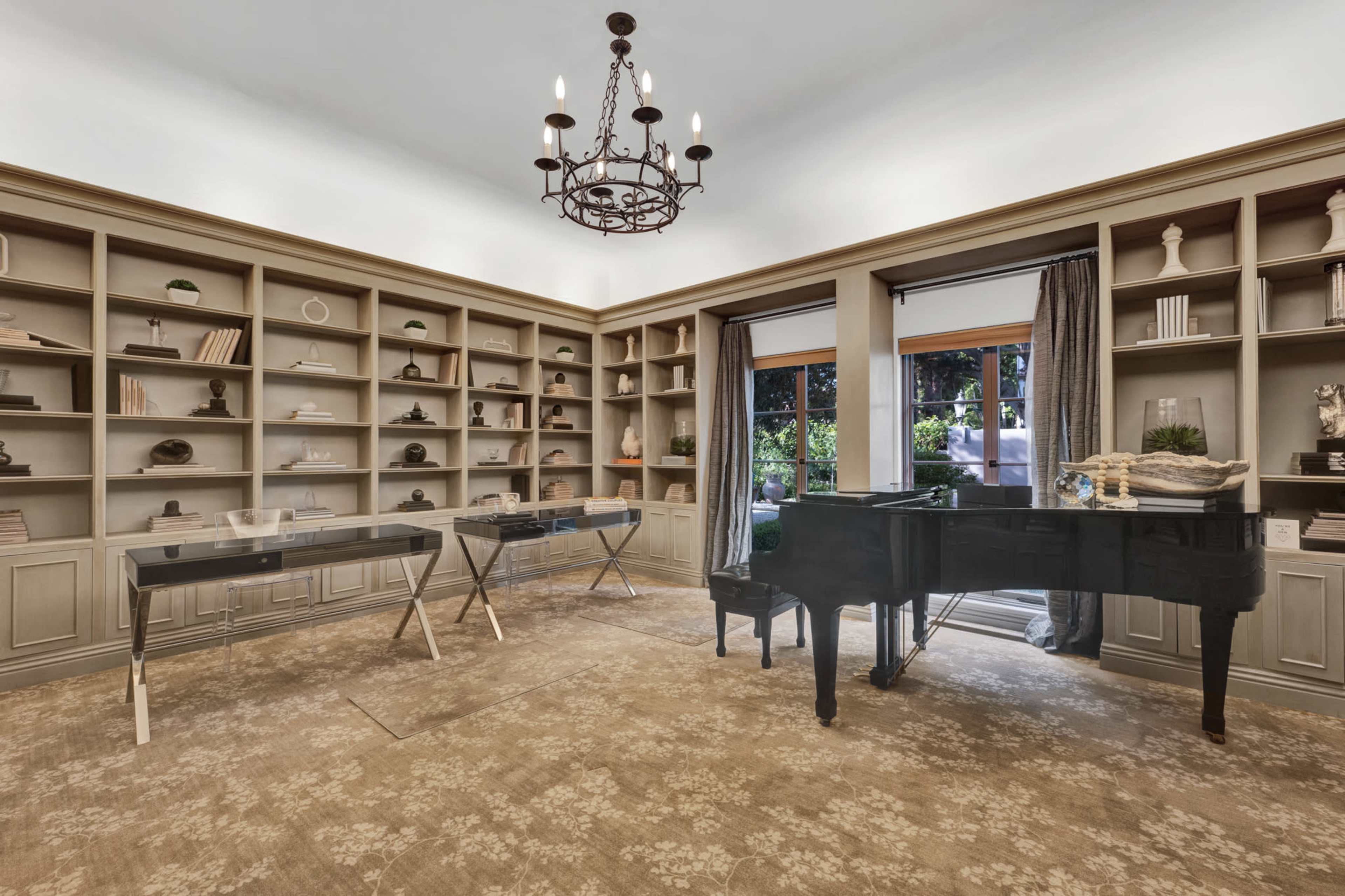 A well-furnished study with a grand piano, two glass desks, and a large bookcase filled with decor items, complemented by windows that provide natural light.