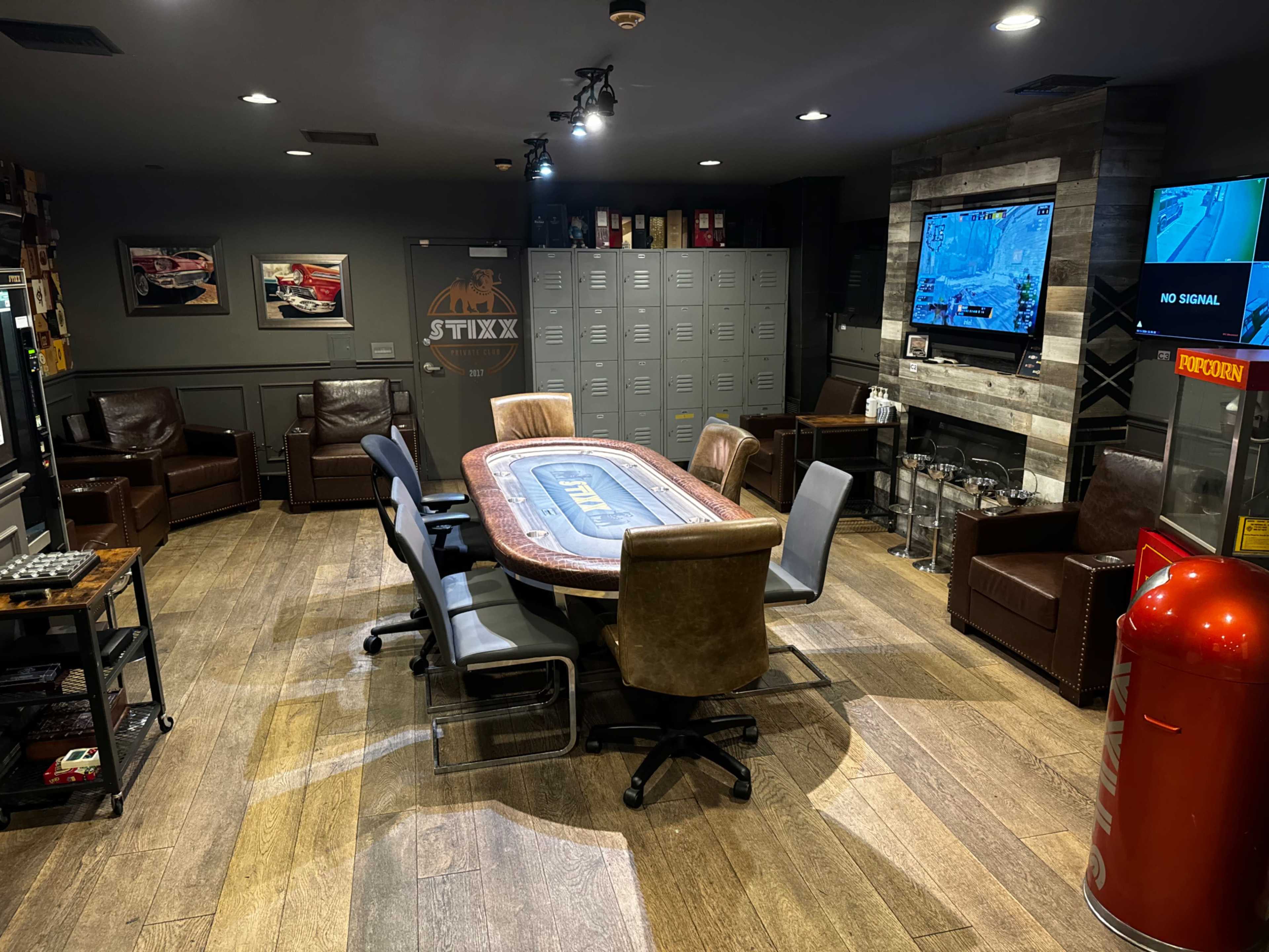 The image shows a cozy game room featuring a large poker table surrounded by chairs, leather armchairs, and a television mounted on the wall, with lockers and a fireplace visible in the background.