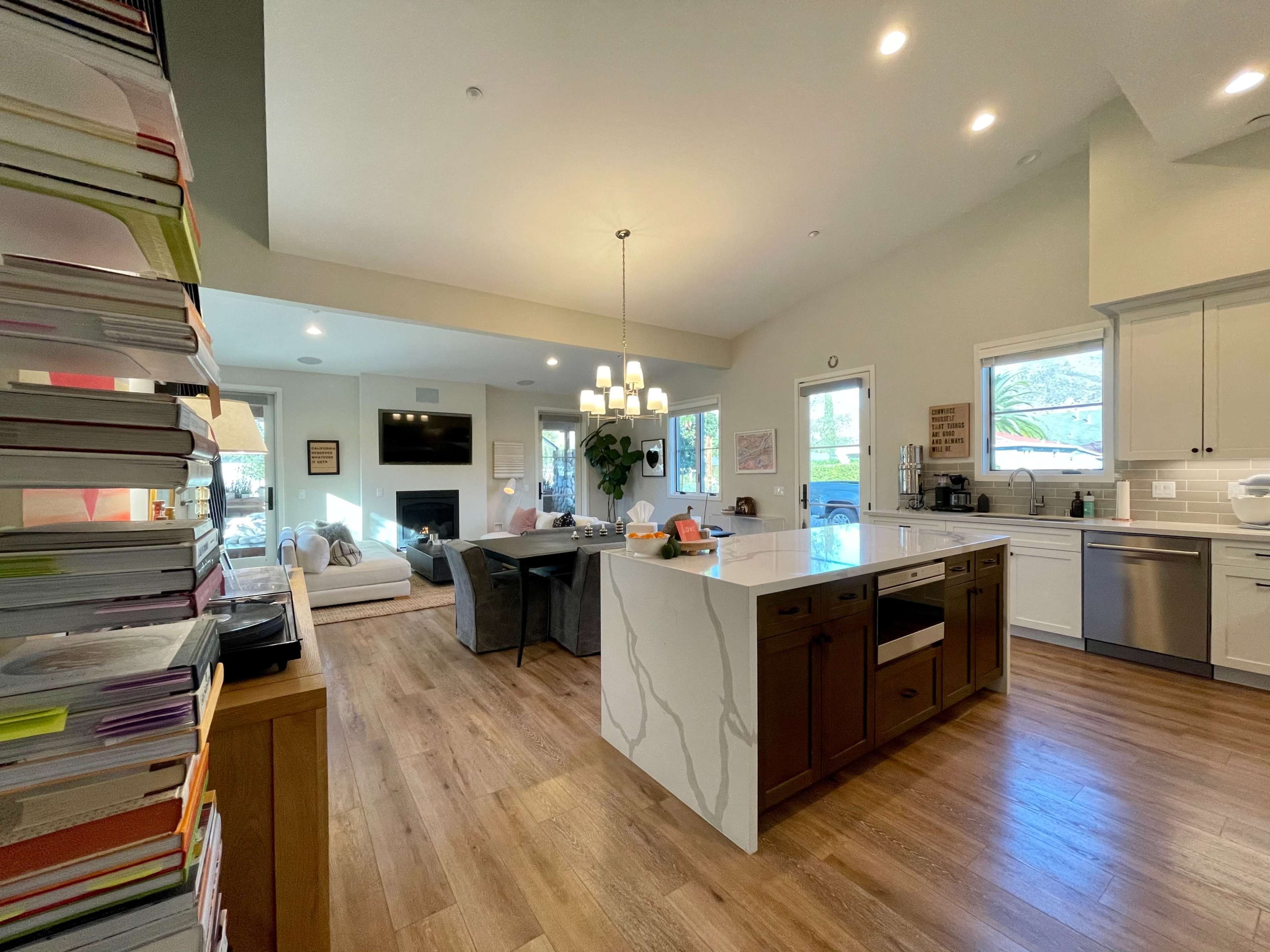 The image shows a modern kitchen and living area with light-colored cabinets, a large island, and a stack of papers on the left.