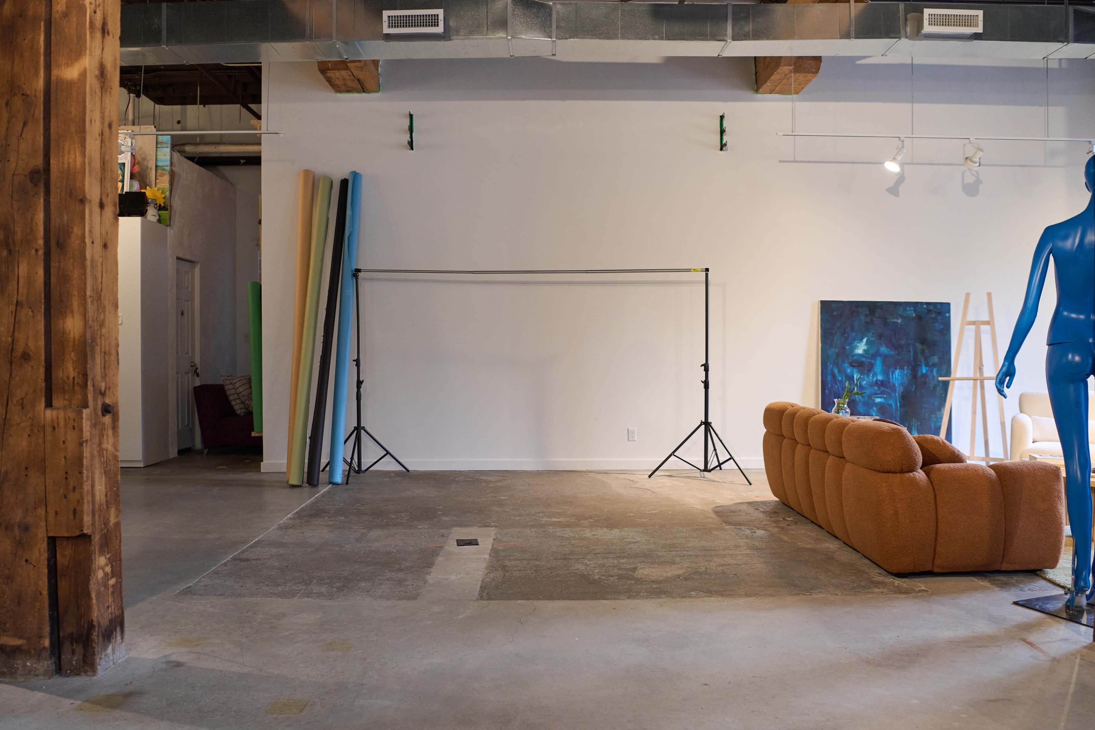 The image shows an empty, well-lit studio space with a large brown sofa, a blue mannequin, and several rolls of backdrop material against the wall.
