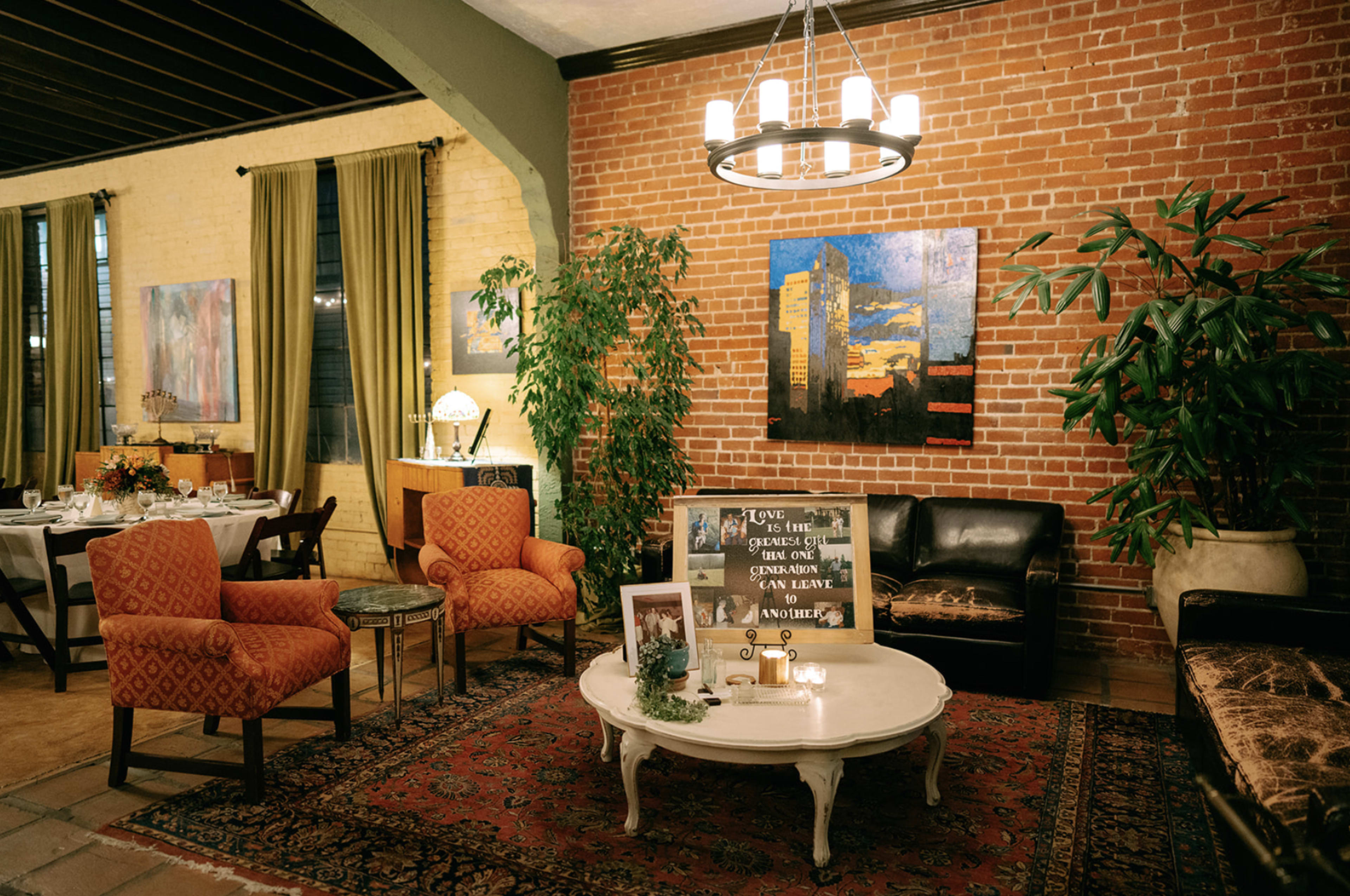 A cozy dining area with brick walls, large paintings, patterned armchairs, a coffee table, and indoor plants.