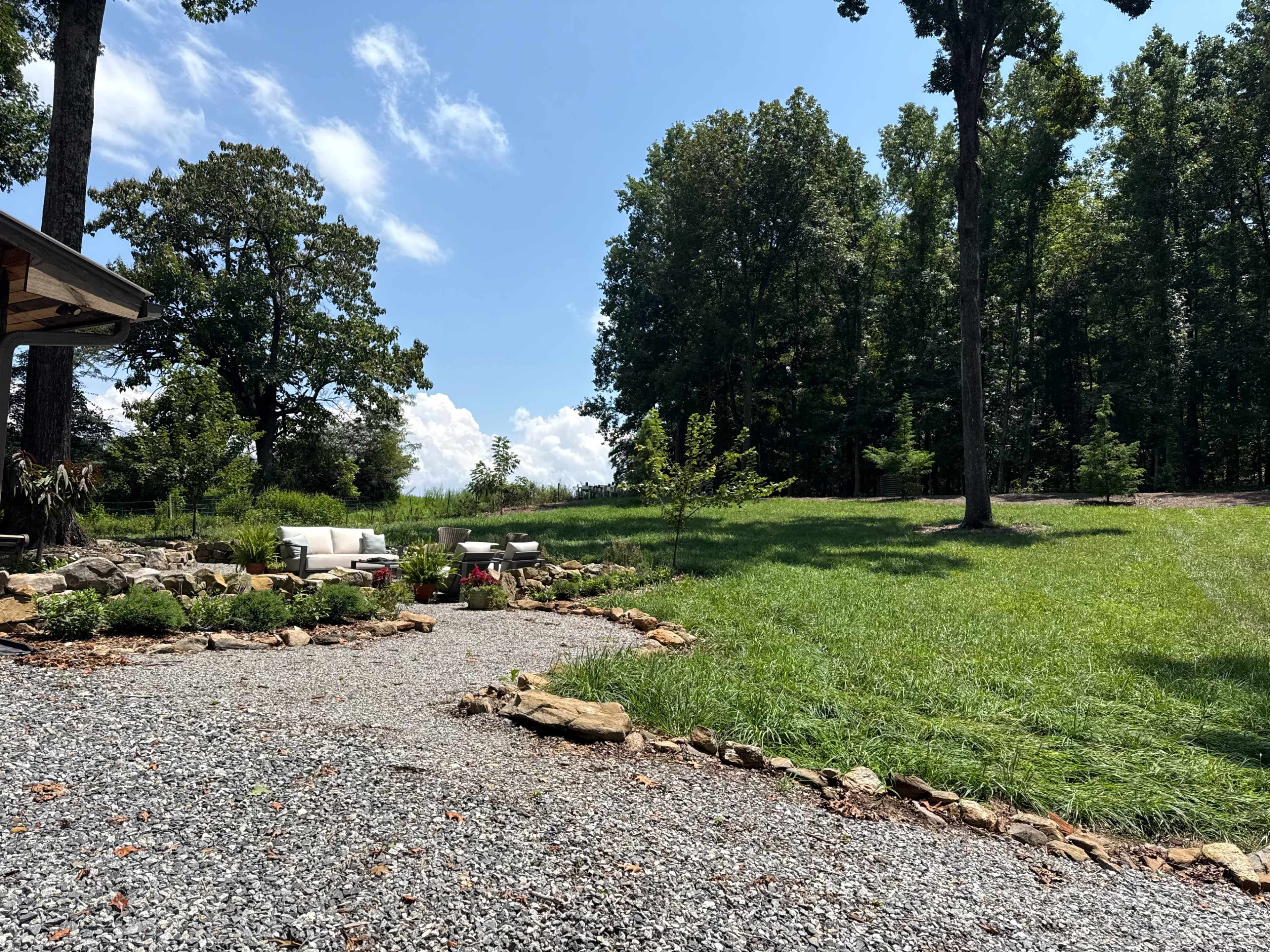 The image shows a gravel path leading through a landscaped area featuring stone borders, with seating arrangements and a grassy field surrounded by trees in the background.