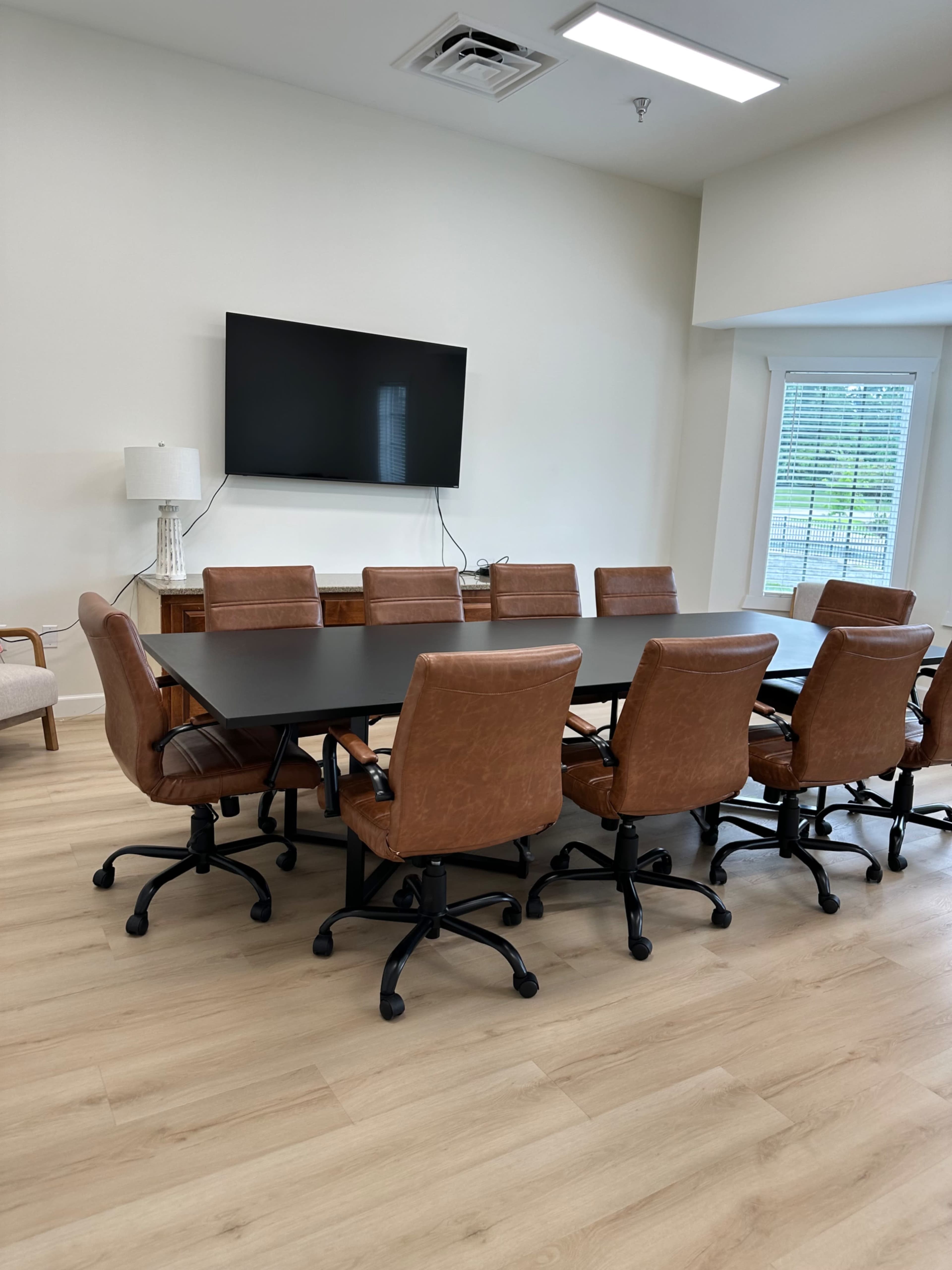A conference room features a large table surrounded by eight brown leather chairs, with a television mounted on the wall and a window providing natural light.
