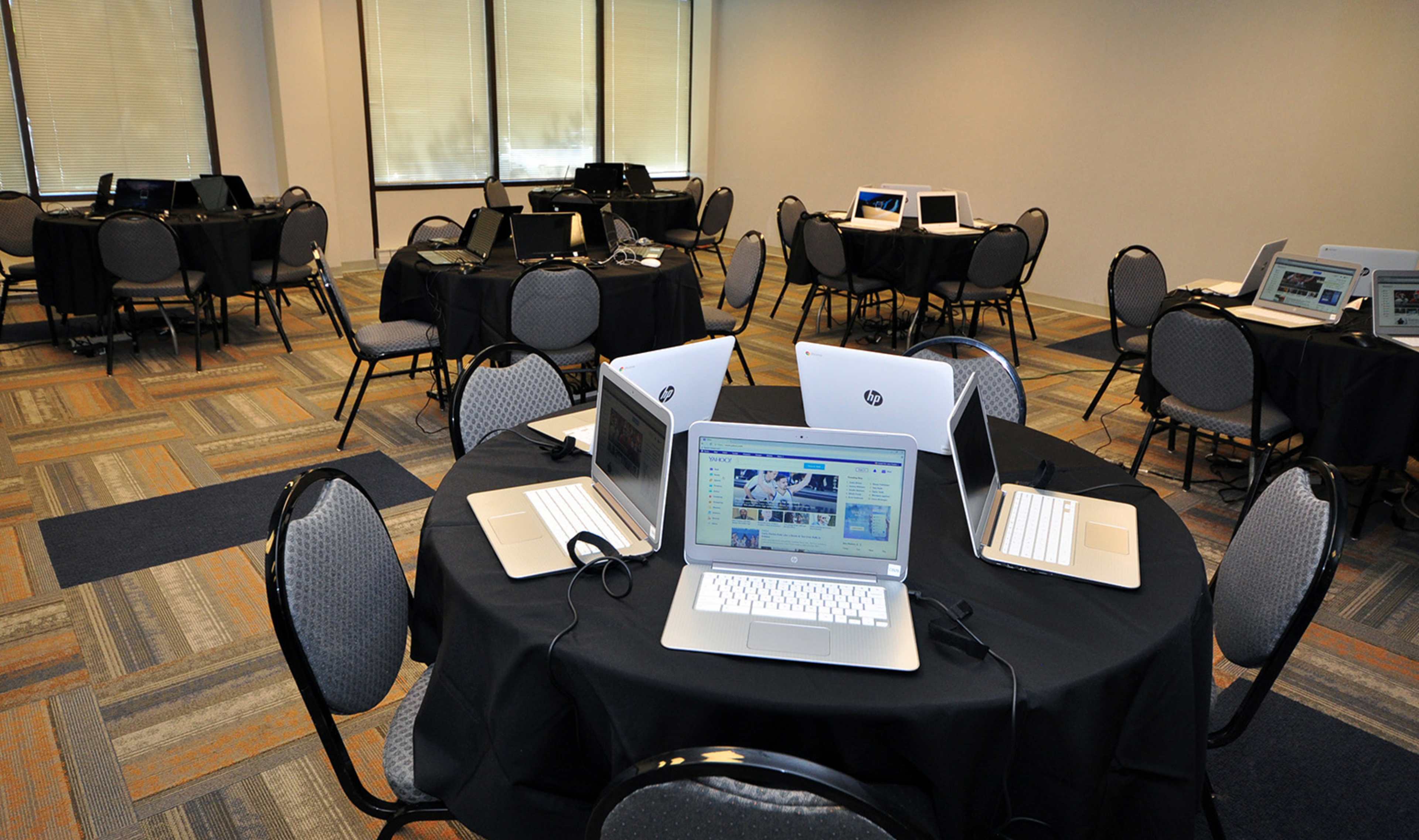 A conference room features several tables with laptops arranged on black tablecloths in a well-lit setting.