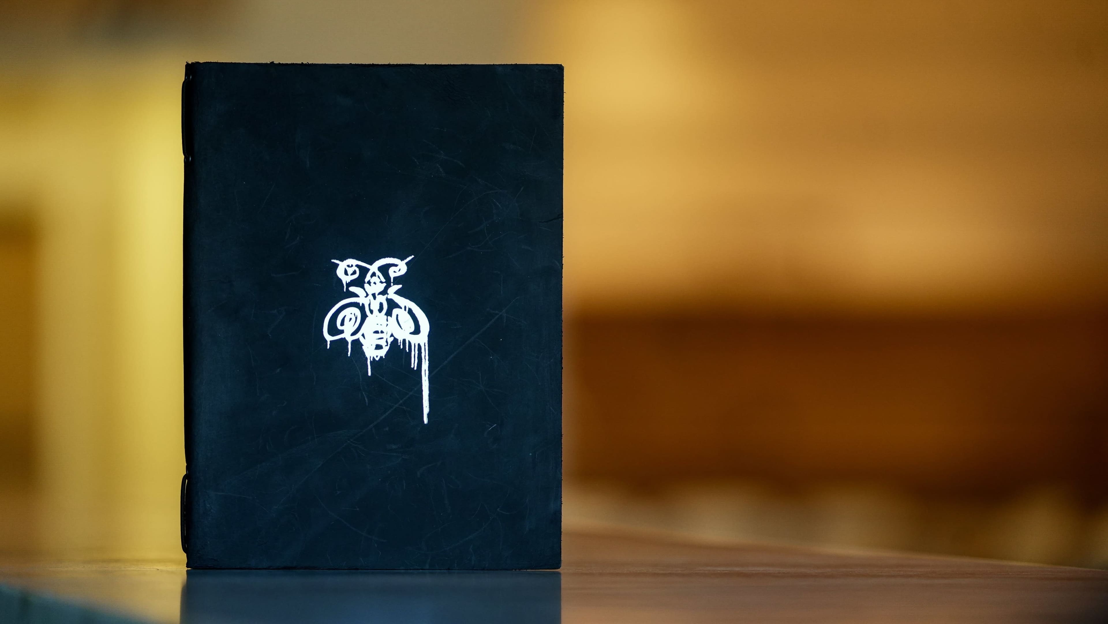 A black notebook with a white design on the cover is placed on a wooden surface, slightly out of focus against a blurred background.